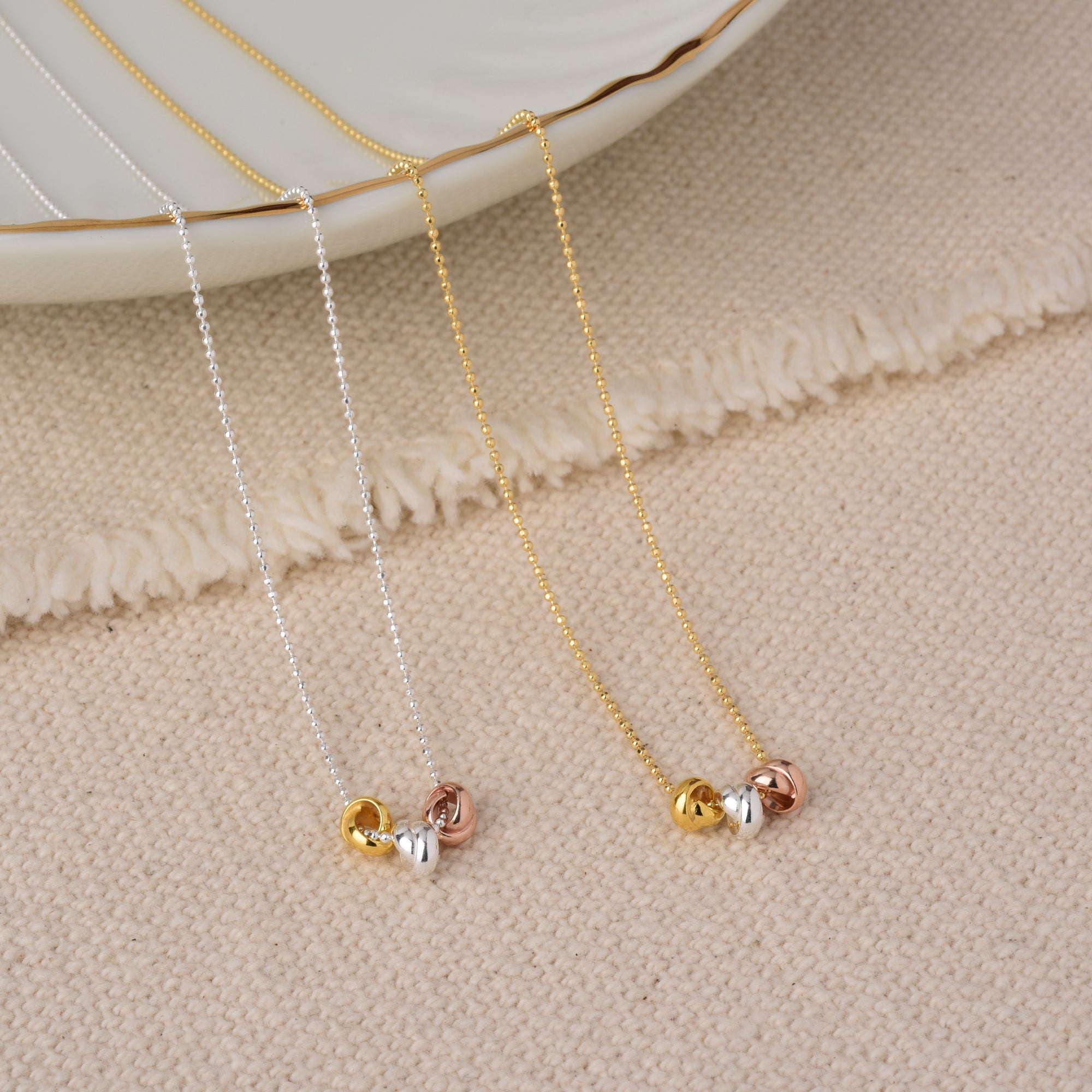 Two delicate necklaces, one silver and one gold, each with three small intertwined rings in gold, silver, and rose gold, displayed on a textured beige fabric next to a decorative white dish.
