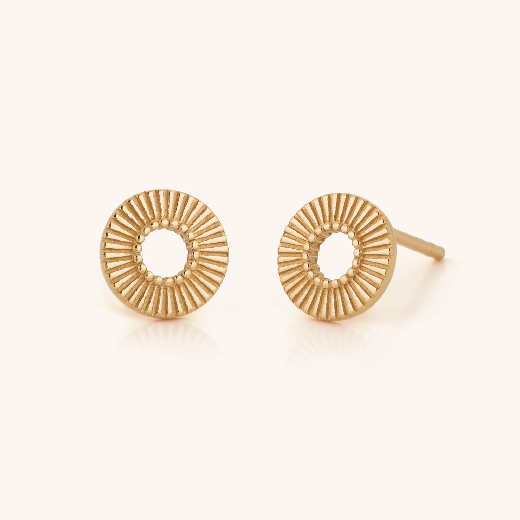 The Solar Circle Earrings are gold studs shaped like small sunbursts, featuring textured ridges radiating from a central open hole for celestial charm.