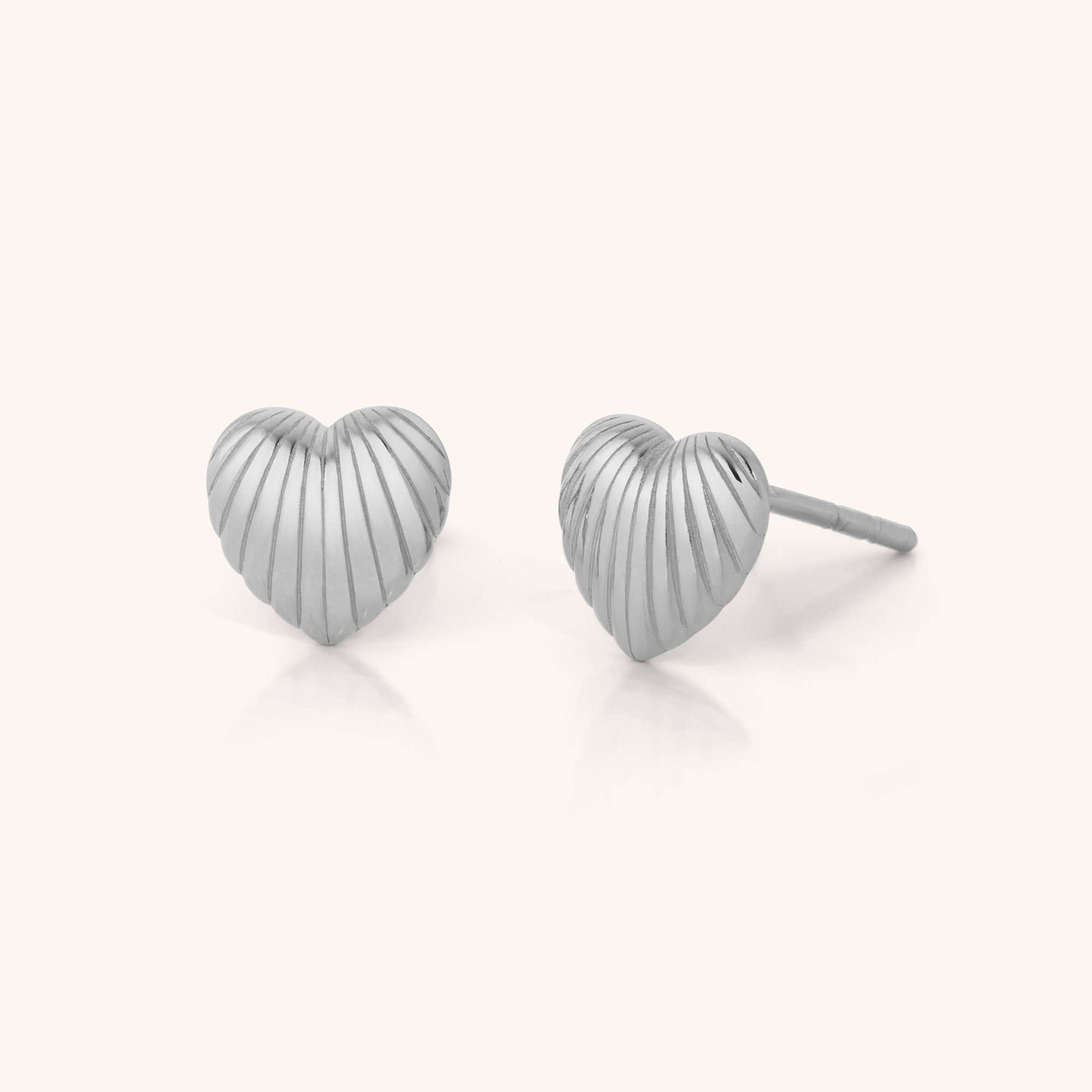 The Heart Beam Stud Earrings—silver, heart-shaped studs with a ridged texture—add elegance and vibrancy to any outfit, shown against a simple, light background.