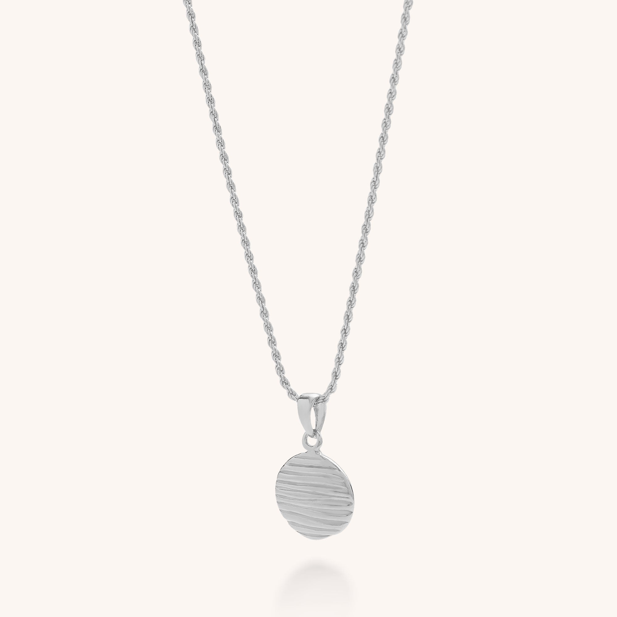 The Wave Disc Necklace features a silver adjustable rope chain and a round pendant with textured horizontal wavy lines, offering modern elegance against a plain light background.