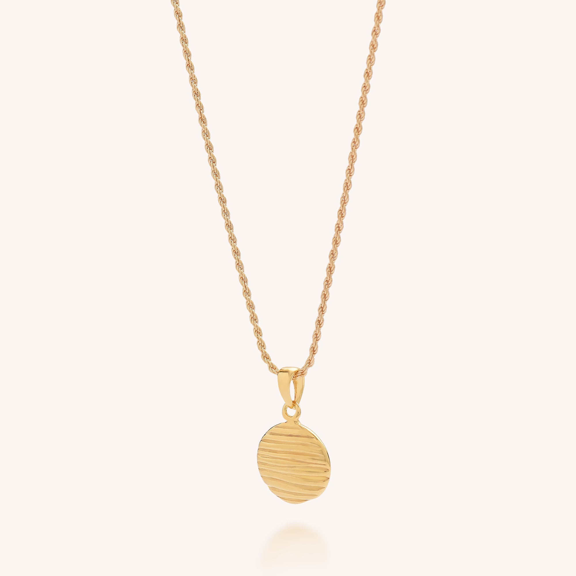 The Wave Disc Necklace features a round, textured gold pendant with wavy lines for a modern elegant look, displayed on a light background.