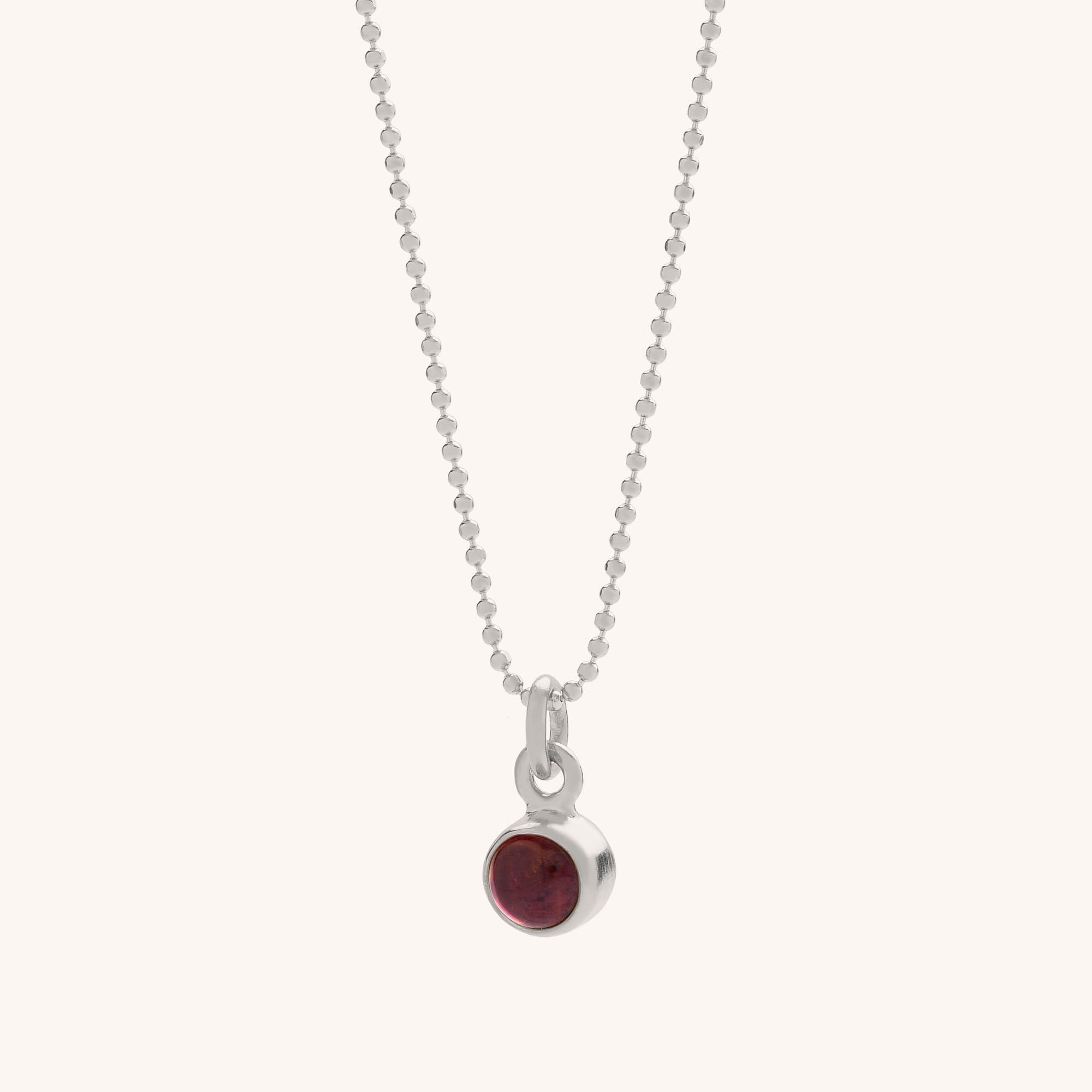 The Gem Dot Birthstone Necklace January features a delicate silver chain with a small round Garnet pendant in a simple silver bezel, an elegant piece believed to offer healing properties.