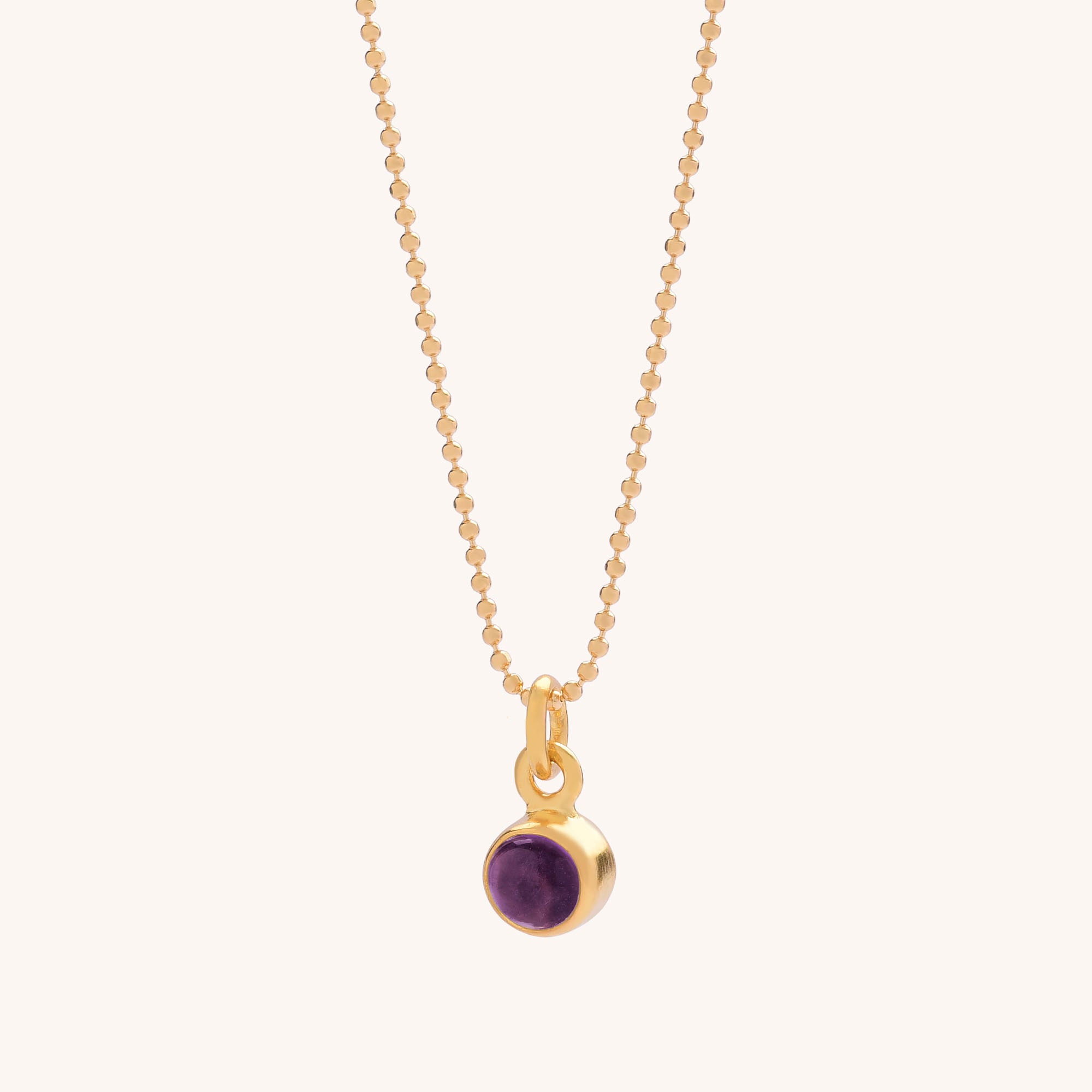 The Gem Dot Birthstone Necklace February is a delicate gold chain featuring a small round amethyst pendant in a gold setting, elegantly displayed on a light background—an elegant and calming birthstone accessory.