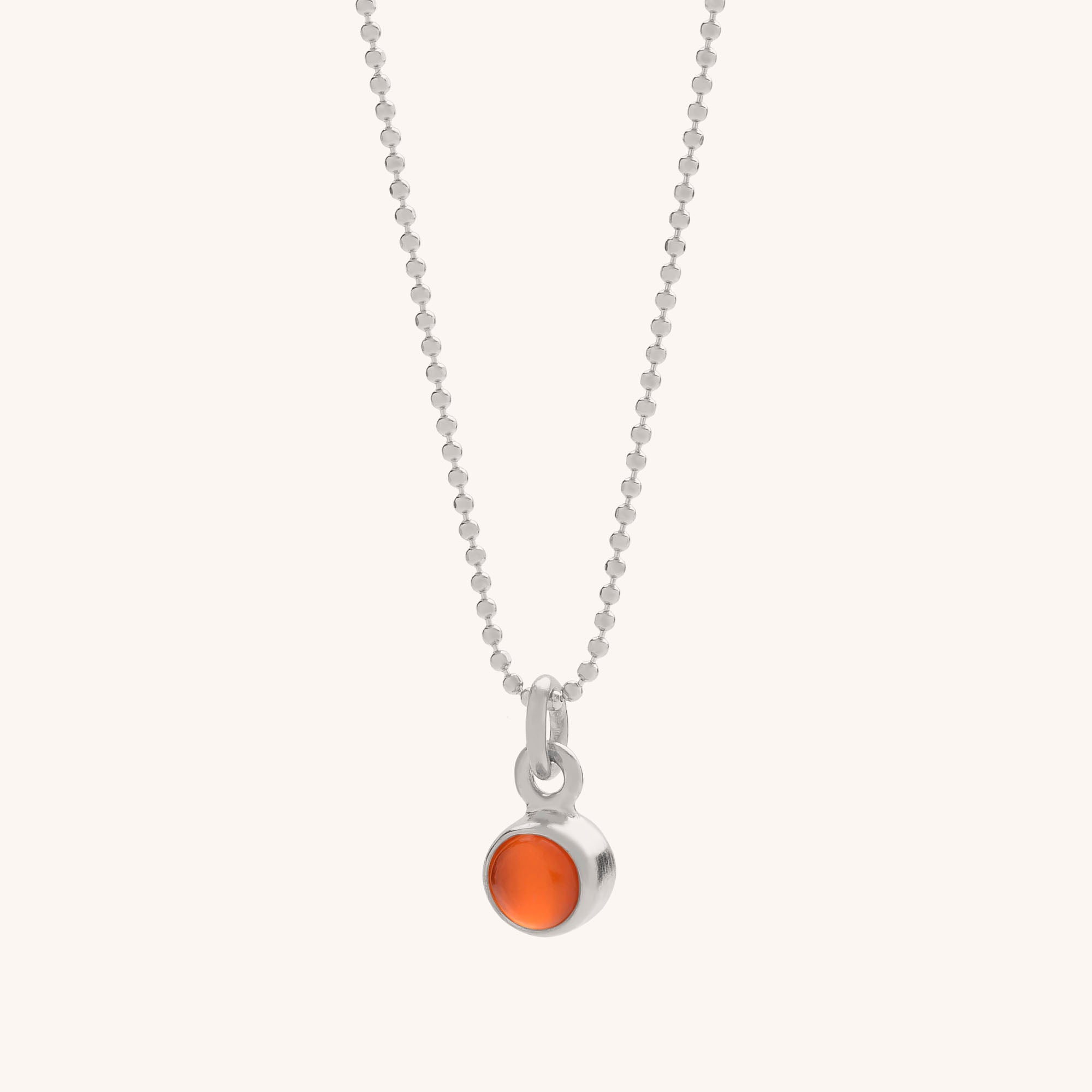 The Gem Dot Birthstone Necklace July features a delicate silver chain with a small, round Carnelian pendant set in a simple silver bezel against a plain, light-colored background.