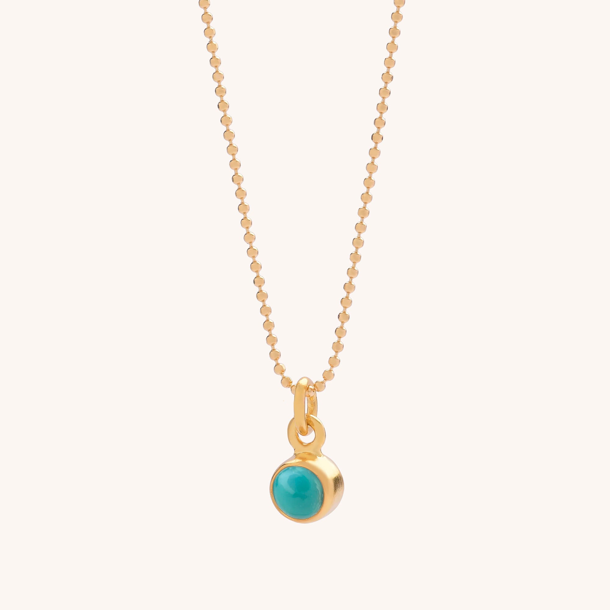 The Gem Dot Birthstone Necklace December features a gold chain with a small, round turquoise gemstone pendant in a gold bezel, elegantly displayed on a light background and believed to offer protection.