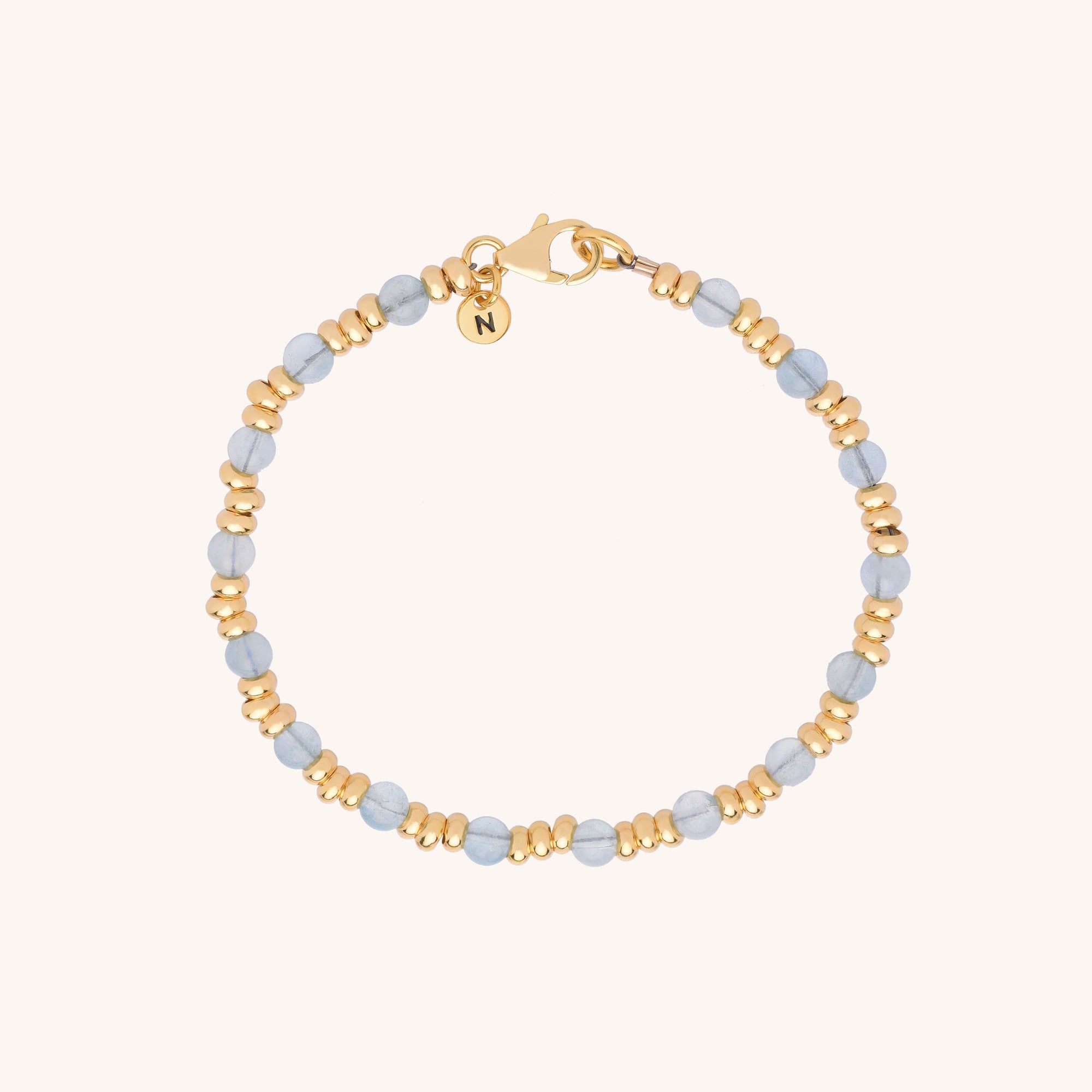 The Gem Bracelet March Birthstone features alternating small gold and aquamarine beads, a gold clasp, and a round charm engraved with N, elegantly displayed on a plain, light background.