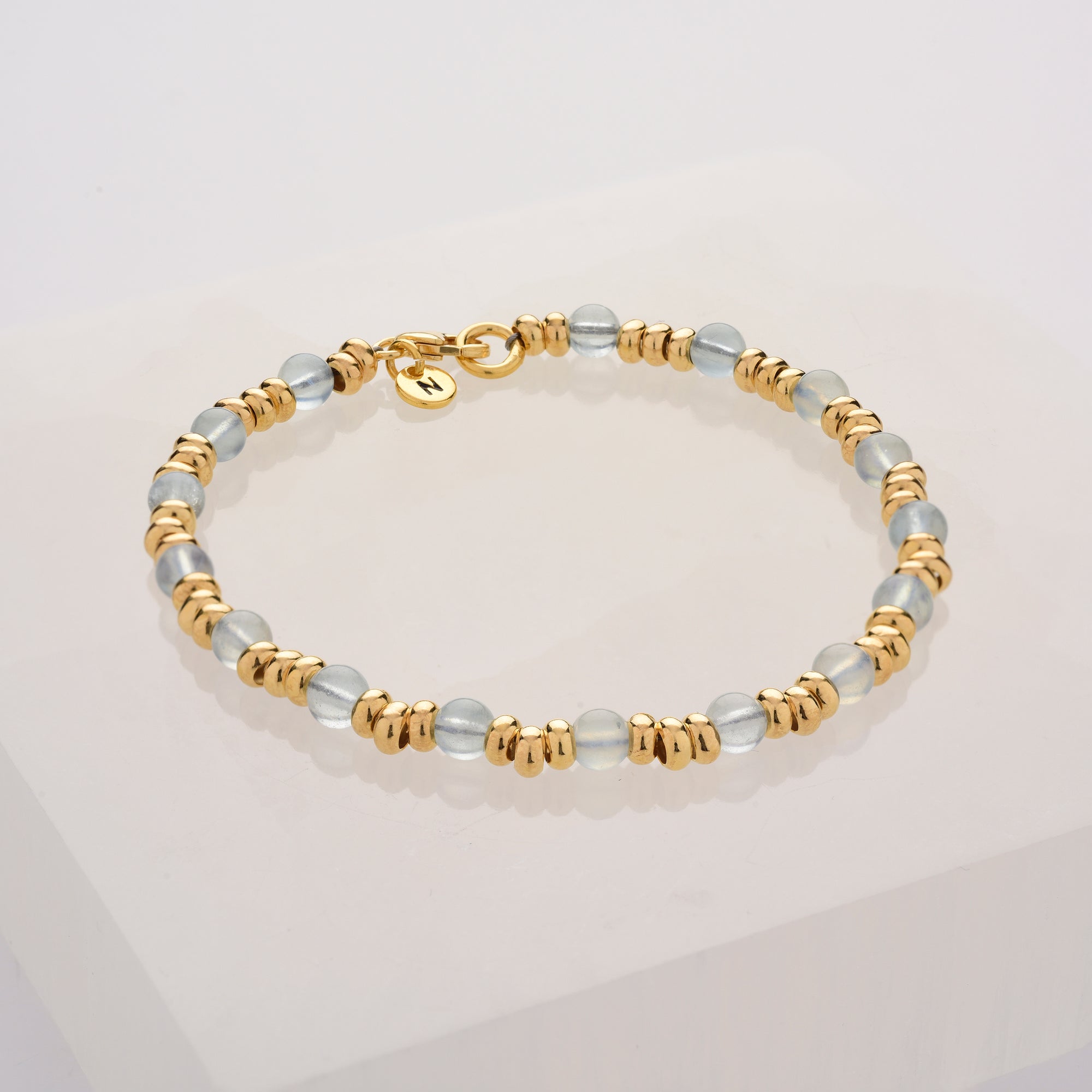 The Gem Bracelet March Birthstone features alternating gold-toned beads and translucent aquamarine-like beads, arranged in a circle on a white background with a small gold charm by the clasp—a refined jewellery piece.