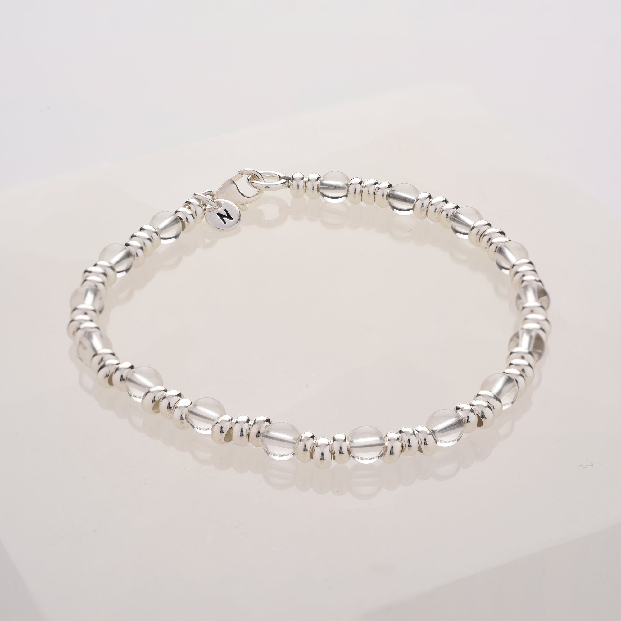 The Gem Bracelet March Birthstone features alternating shiny and matte silver oval beads with a delicate metallic “N” charm, elegantly displayed on a white surface.