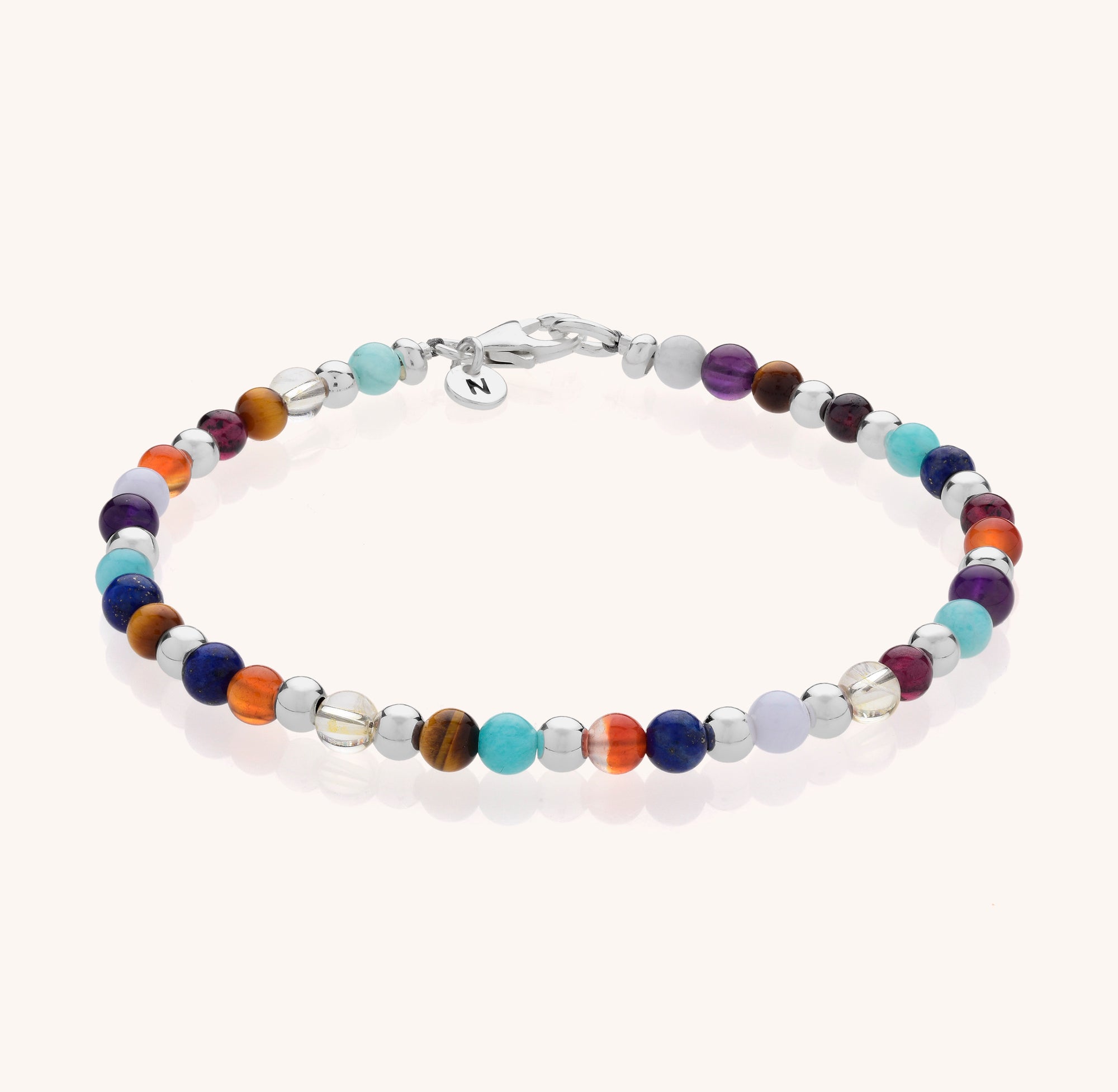 The Multi Gemstone Bracelet showcases small round beads in blue, orange, purple, brown, and clear natural gemstones with a silver clasp and round tag, beautifully displayed on a light background.