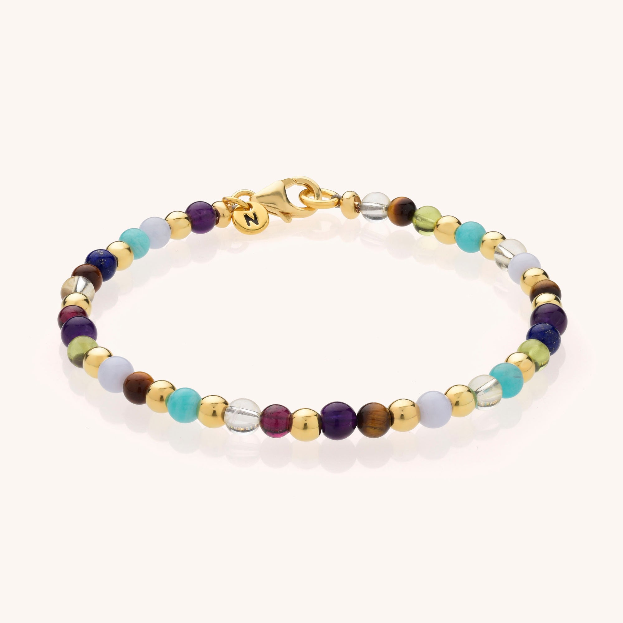The Multi Gemstone Bracelet features alternating gold, turquoise, blue, purple, brown, clear, and white natural gemstone beads with a gold clasp and a small gold charm engraved with the letter N.