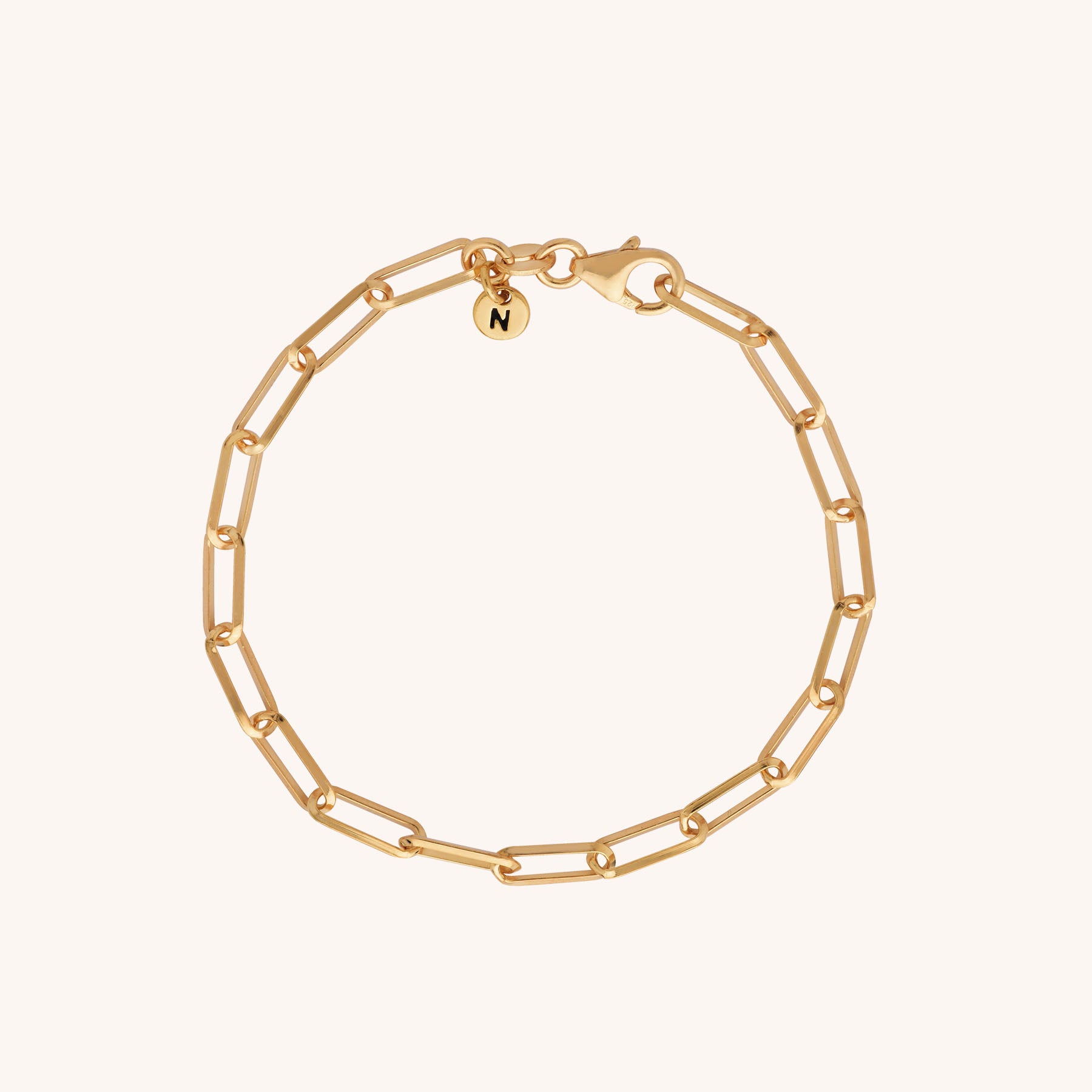 The Triangular Paperclip Bracelet is a sustainable gold accessory featuring elongated oval links and a small round charm engraved with “N,” shown on a plain light background.