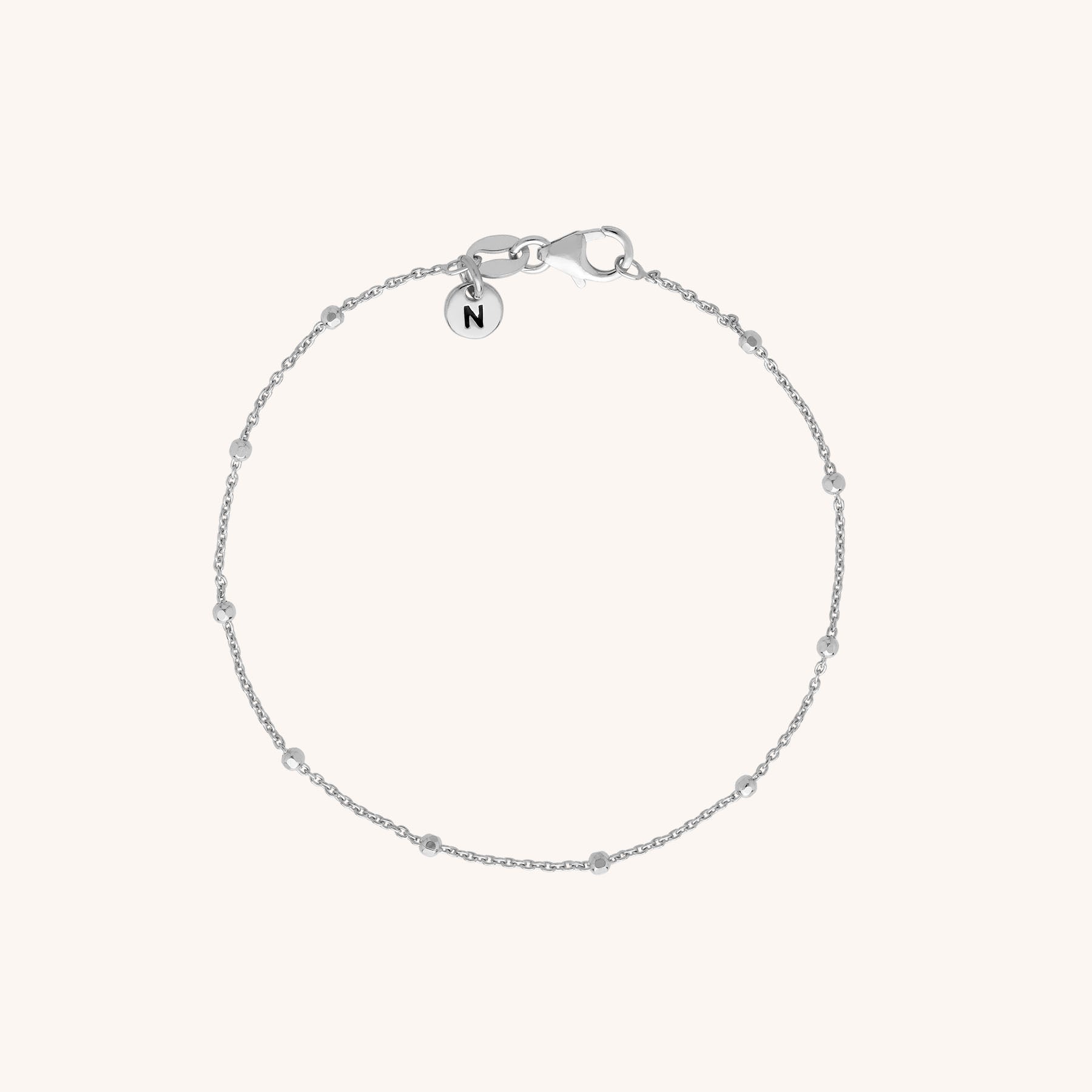 The Satellite Bracelet is a delicate eco-silver piece with evenly spaced small beads and a round N-engraved charm near the clasp, shown on a light background.