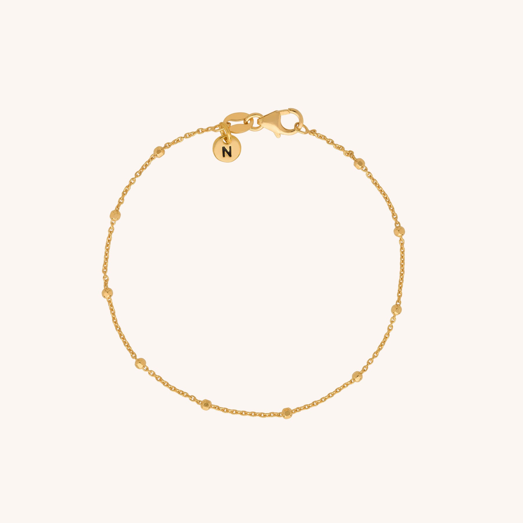The Satellite Bracelet is a delicate gold chain with evenly spaced small beads and a round charm engraved with the letter N near the clasp—a stunning piece of sustainable jewellery.