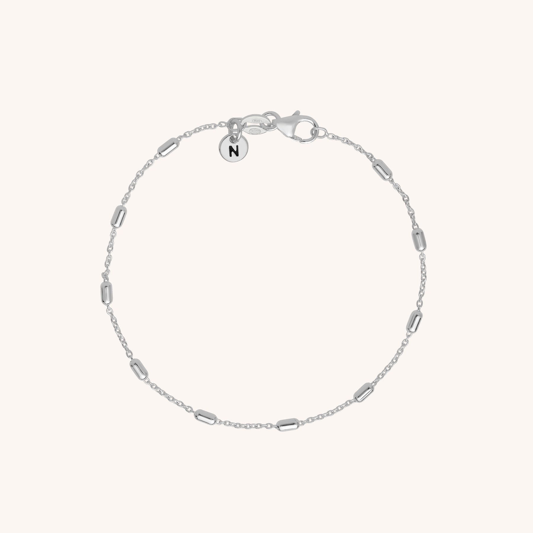 The Tubular Satellite Bracelet features small cylindrical beads on an eco silver chain, a lobster clasp, and a round charm engraved with the letter N, shown on a plain light background.