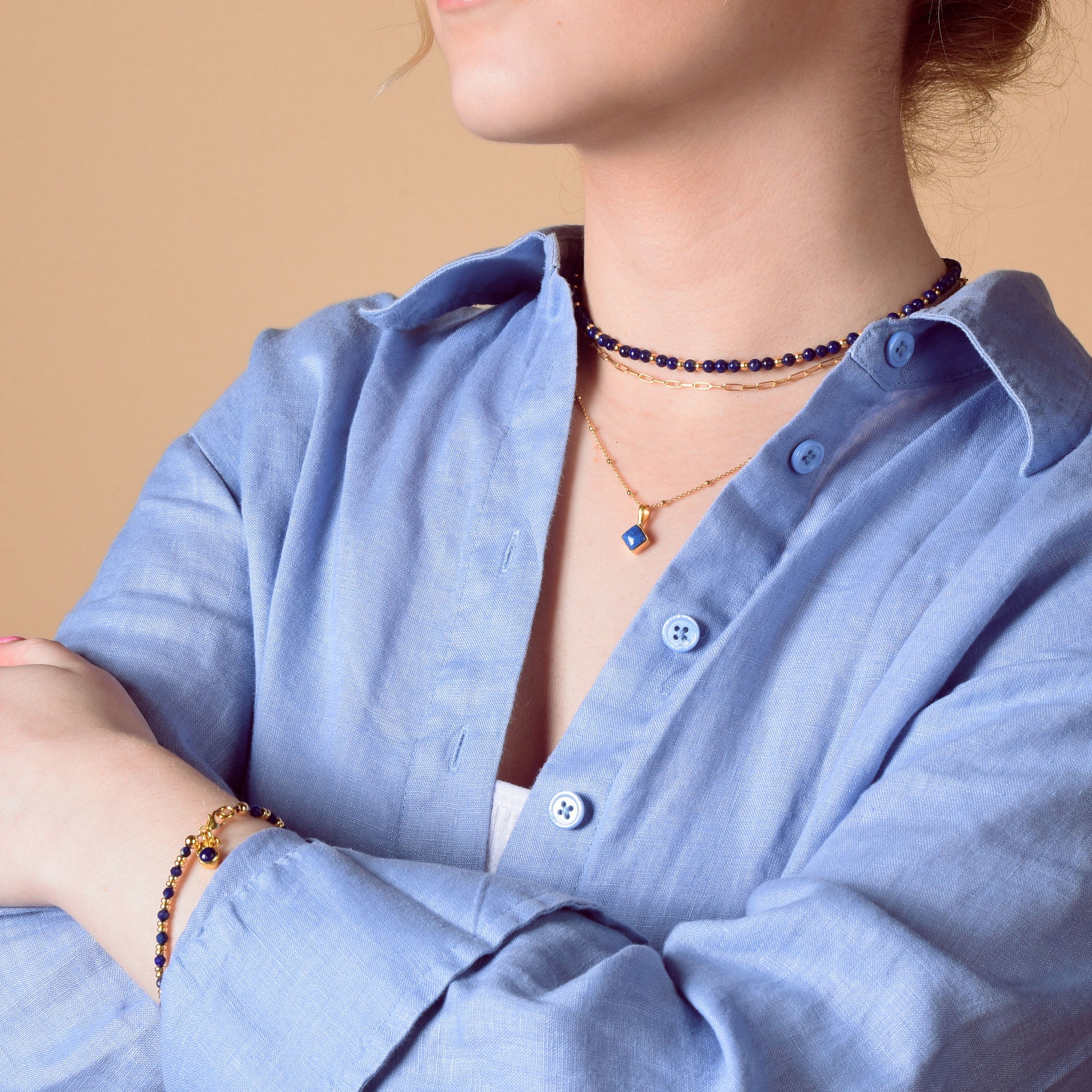 A woman in a light blue button-up shirt wears layered gold and dark blue beaded necklaces, including the eco-friendly Jewel Bead Birthstone Necklace - September, plus a matching bracelet, standing with arms folded against a beige background.