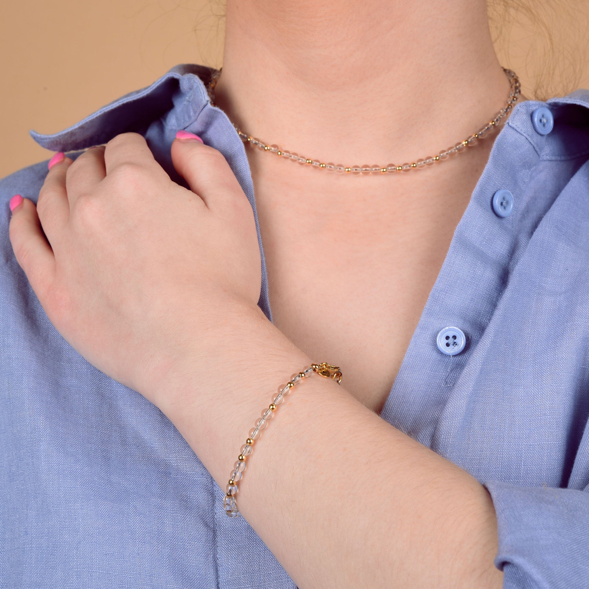 A person in a light blue button-up shirt showcases the handmade Jewel Bead Birthstone Necklace - April, crafted in the UK, along with a delicate gold chain bracelet, their hand resting on their chest.