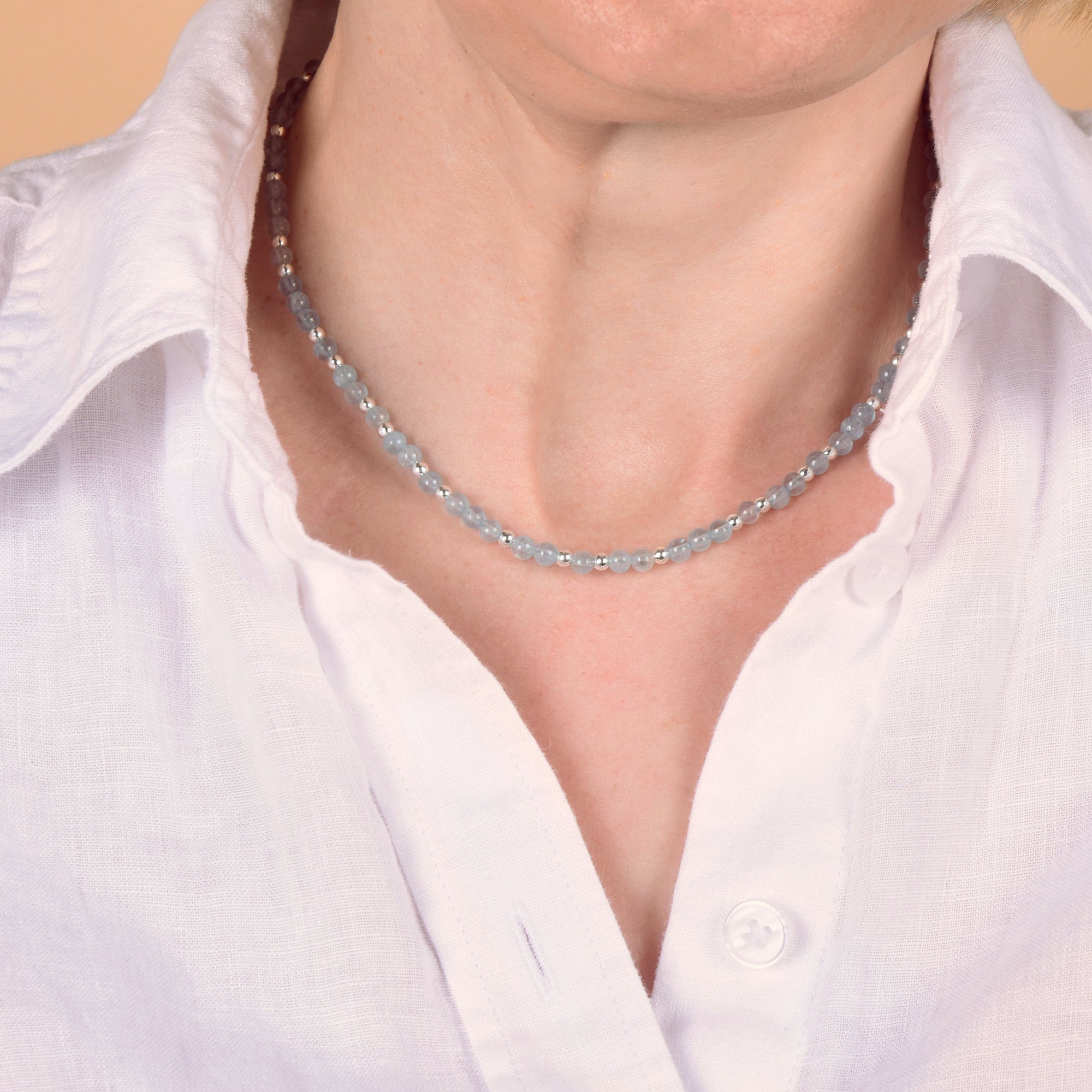 Close-up of a person wearing a white collared shirt and the Jewel Bead Birthstone Necklace - March, featuring delicate aquamarine, light blue, and clear beads—an elegant accessory that highlights the March birthstone. Focus is on the neck and collar area.