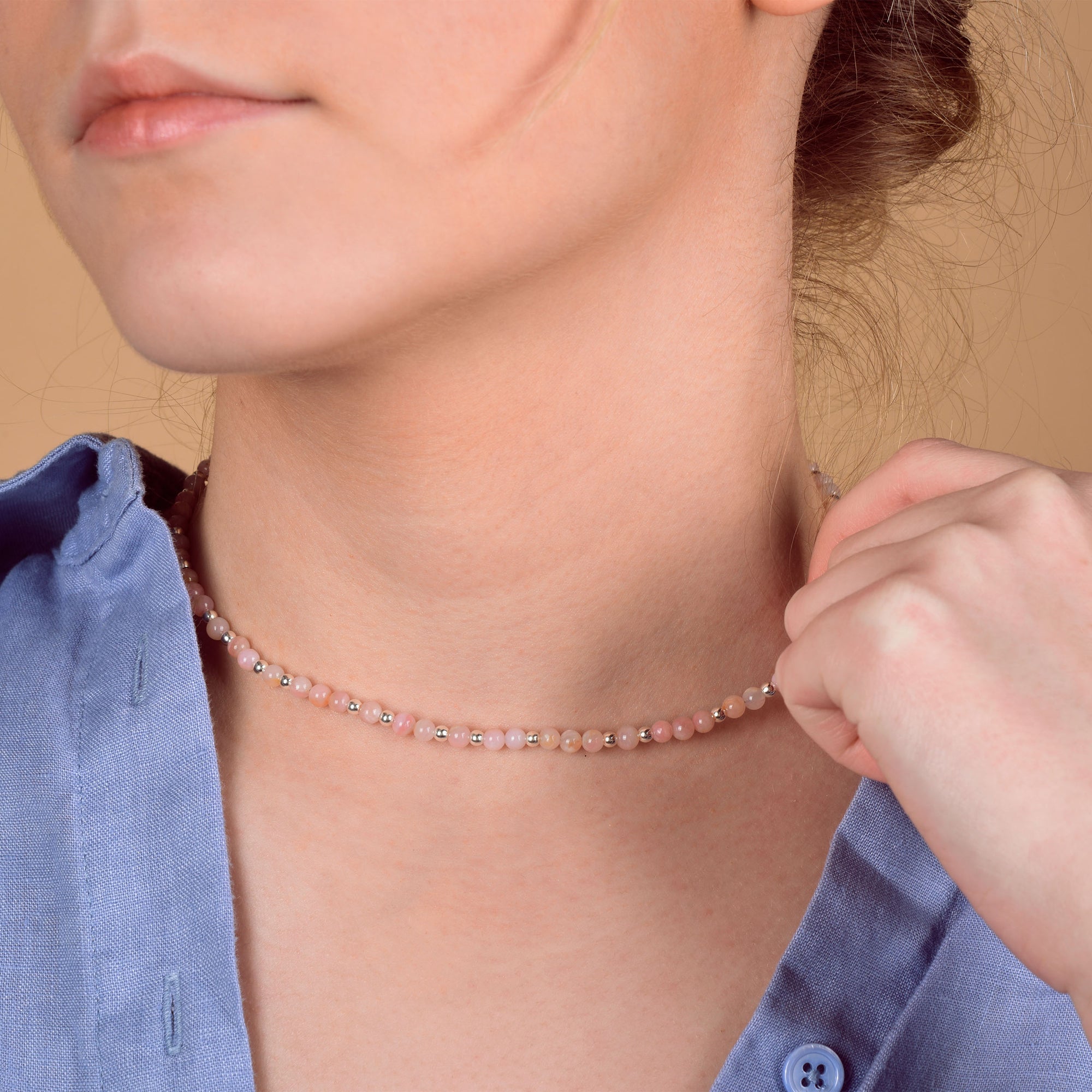 A person in a blue shirt holds the Jewel Bead Birthstone Necklace - October, a delicate Pink Opal beaded choker, around their neck against a soft beige background. Only the lower face and neck are visible.