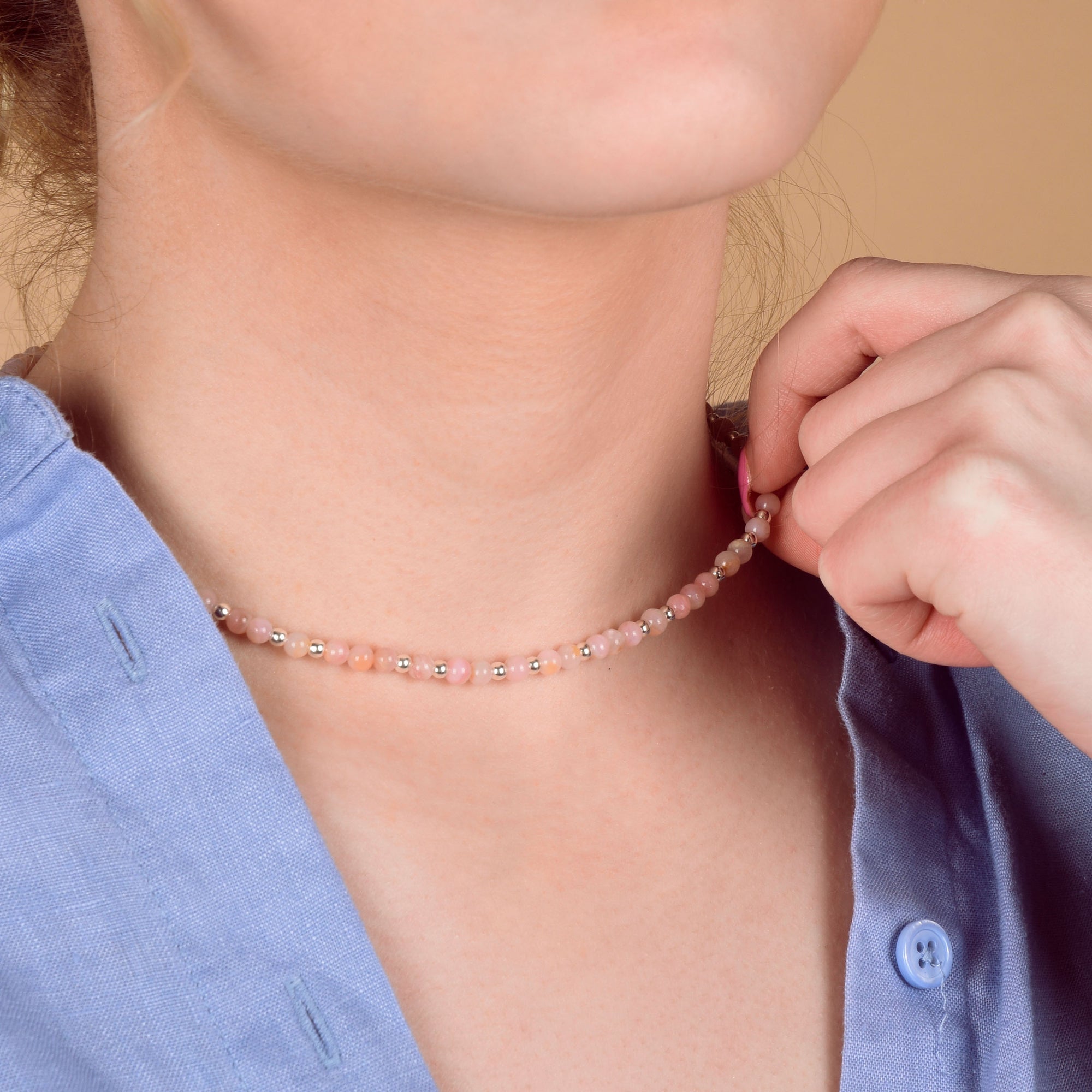 A person in a light blue shirt holds the Jewel Bead Birthstone Necklace - October, a delicate choker with pink and white beads featuring Pink Opal accents, around their neck against a neutral beige background.