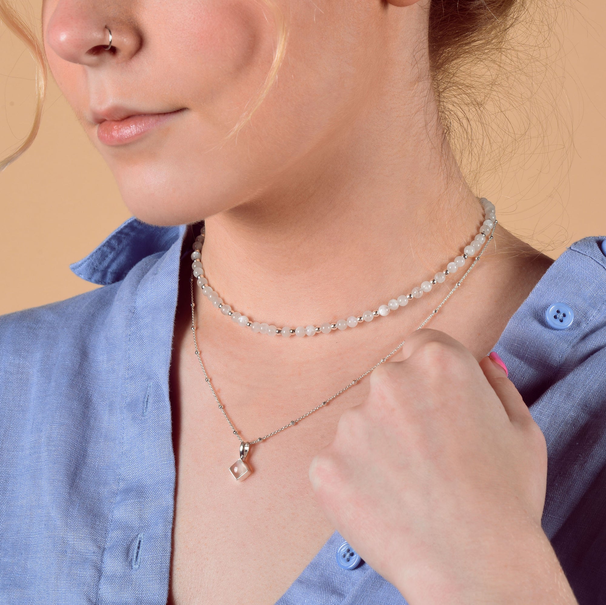 A woman wears a light blue button-up shirt and the Jewel Bead Birthstone Necklace - June, layered with a longer Rainbow Moonstone pendant. She has a nose piercing and light pink nail polish, with her hand near her collar.
