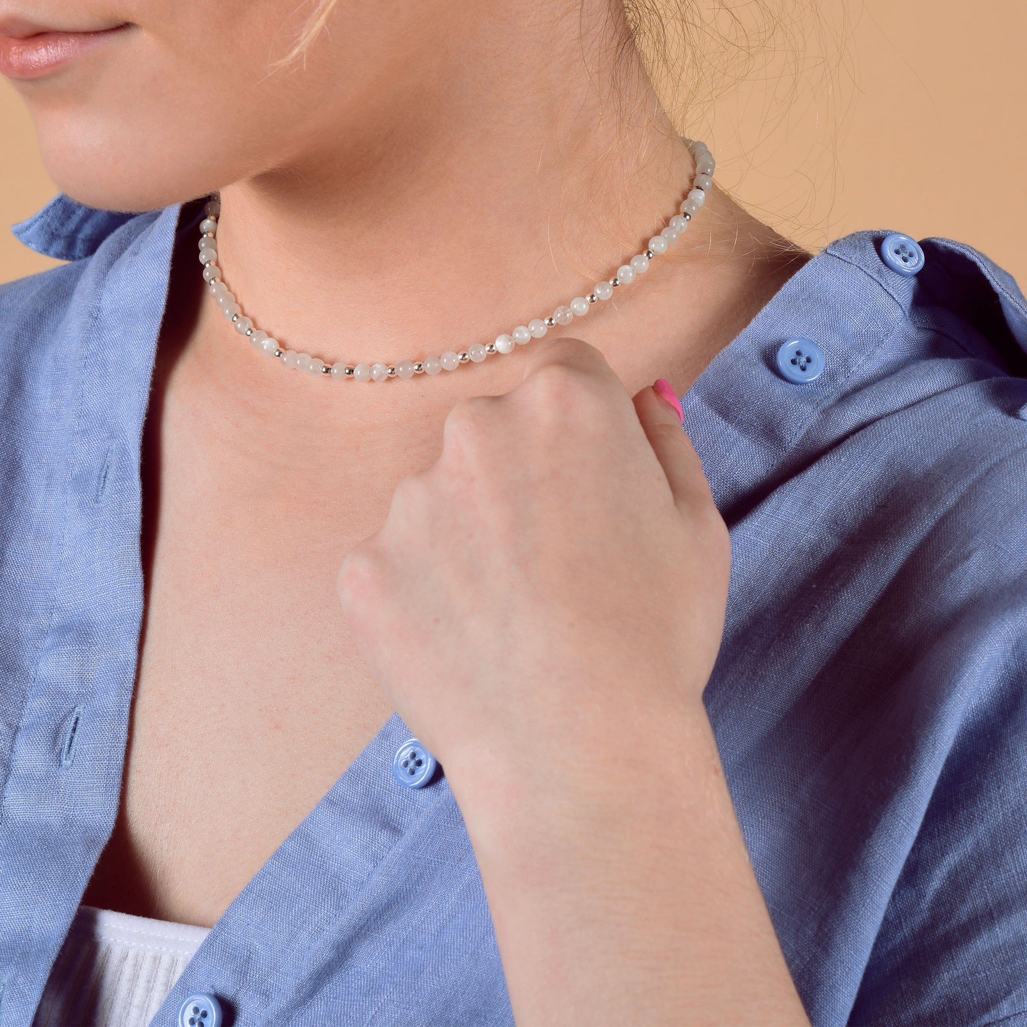 A woman in a light blue button-up shirt showcases the Jewel Bead Birthstone Necklace - June. She holds her pink-painted nails near her collar against a neutral peach background.