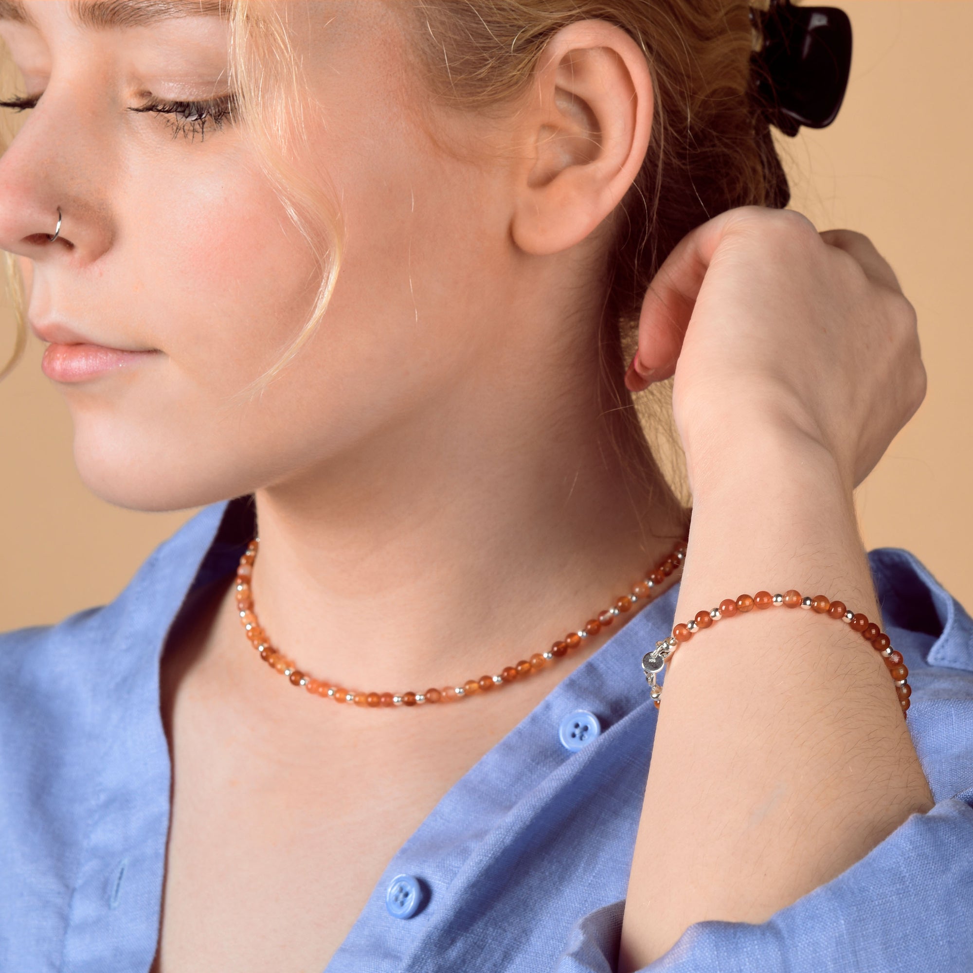 A woman with light hair wears a blue shirt and the Jewel Bead Birthstone Necklace - July, along with a matching bracelet. She gazes to the side, hand near her neck, highlighting the jewelry set against a soft beige background.