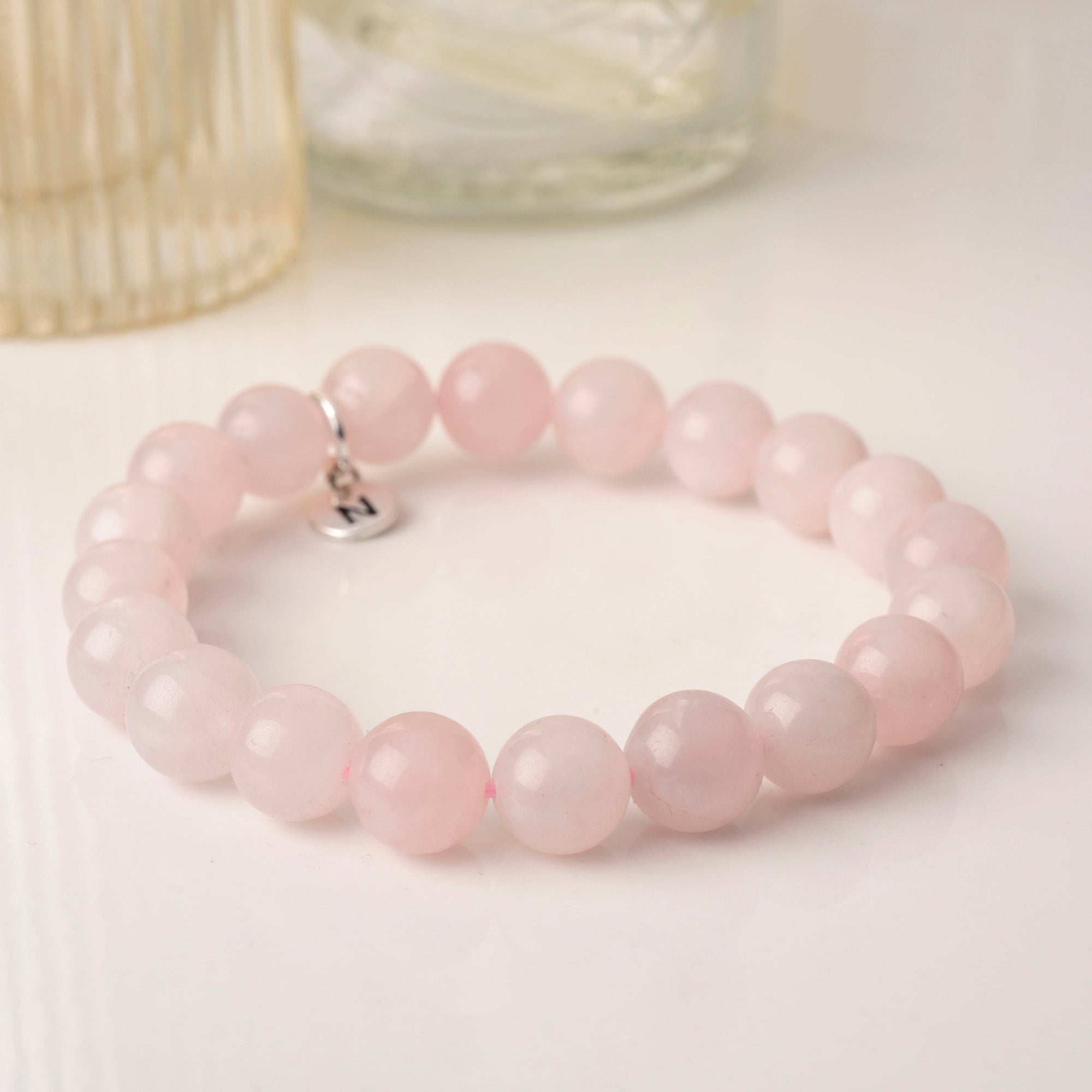 The Affinity Gemstone Bracelet features large round light pink beads and a small silver charm, displayed on a white surface beside glass containers.