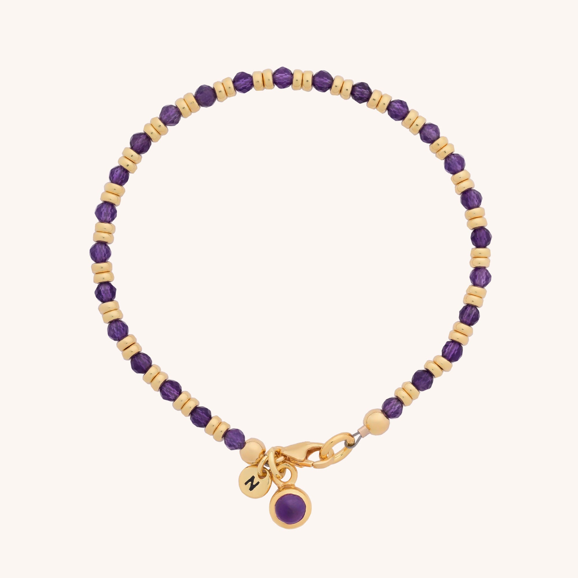 The Linear Birthstone Bracelet is a delicate beaded piece with alternating gold and purple beads, natural gemstones, a gold clasp, and two gold charms—one engraved, one set with a purple stone. Ideal for gemstone jewellery lovers.