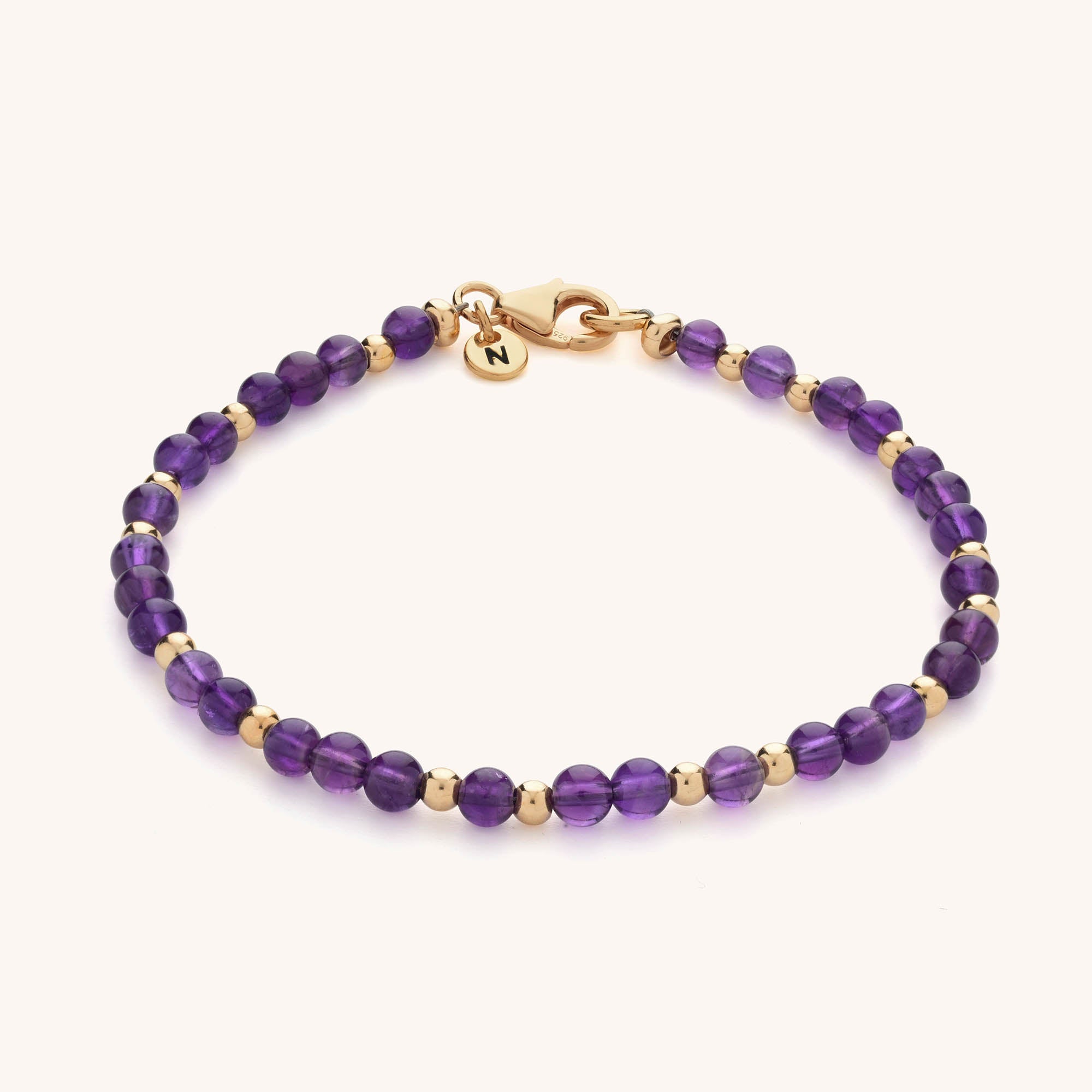 The Jewel Bead Birthstone Bracelet features small round purple natural gemstones alternating with gold beads, finished with a gold clasp and a round gold charm engraved with the letter N.