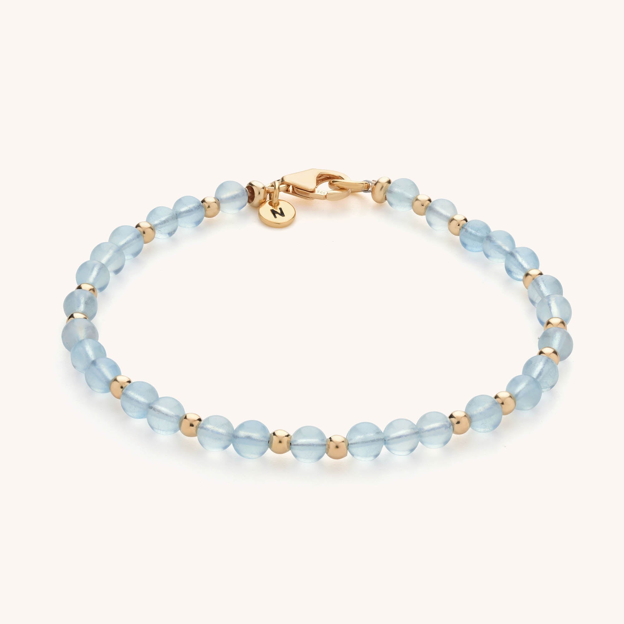 The Jewel Bead Birthstone Bracelet - March showcases round, translucent aquamarine beads separated by gold accents, with a gold clasp and a dainty gold charm engraved with the letter N.