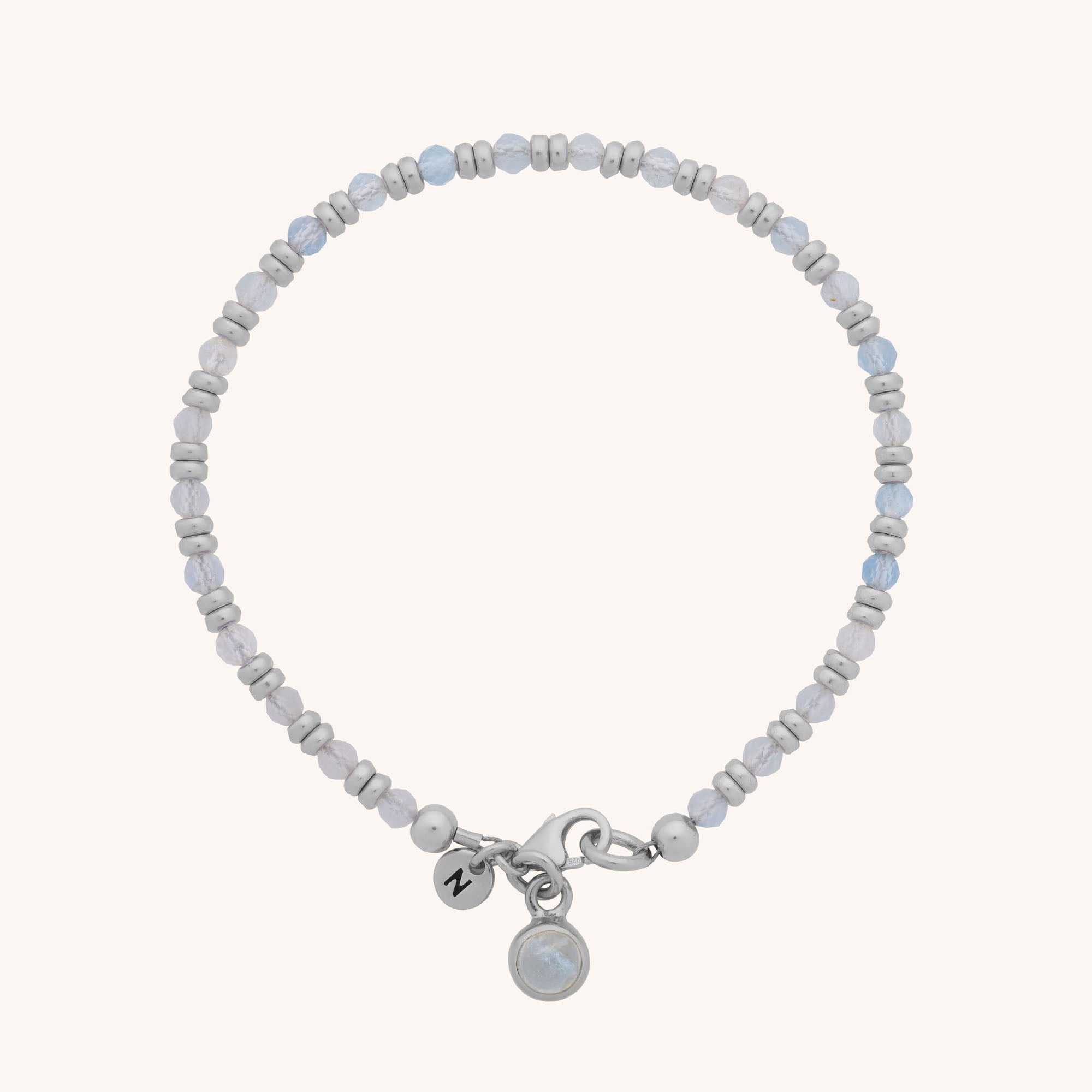 The Linear Birthstone Bracelet features small light grey and pale blue beads, a silver clasp, a round charm with a translucent natural gemstone, and a circular tag engraved with the letter Z for an elegant touch.