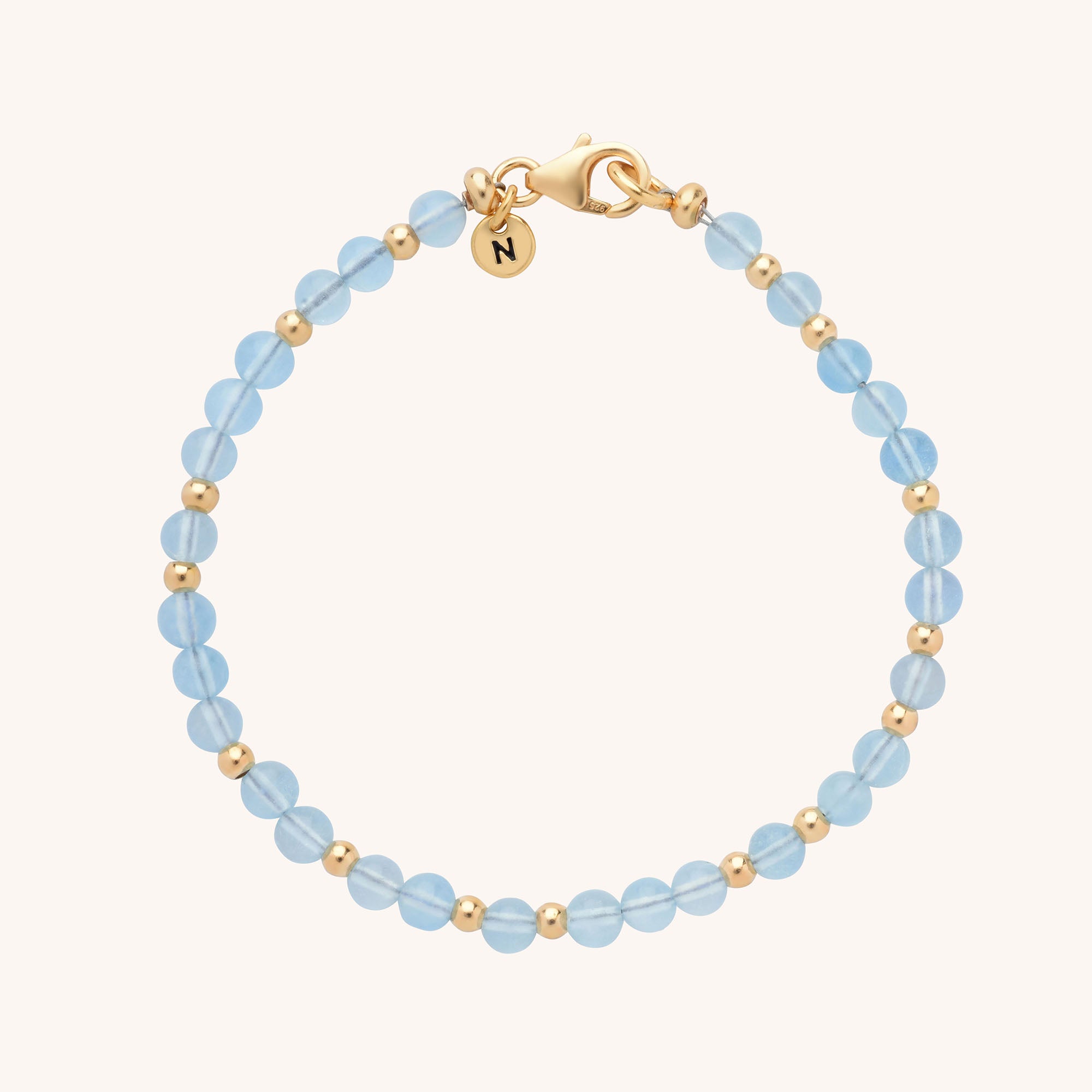 The Jewel Bead Birthstone Bracelet - March features alternating light blue aquamarine gemstones and gold beads, finished with a gold lobster clasp and a round charm engraved with the letter N.