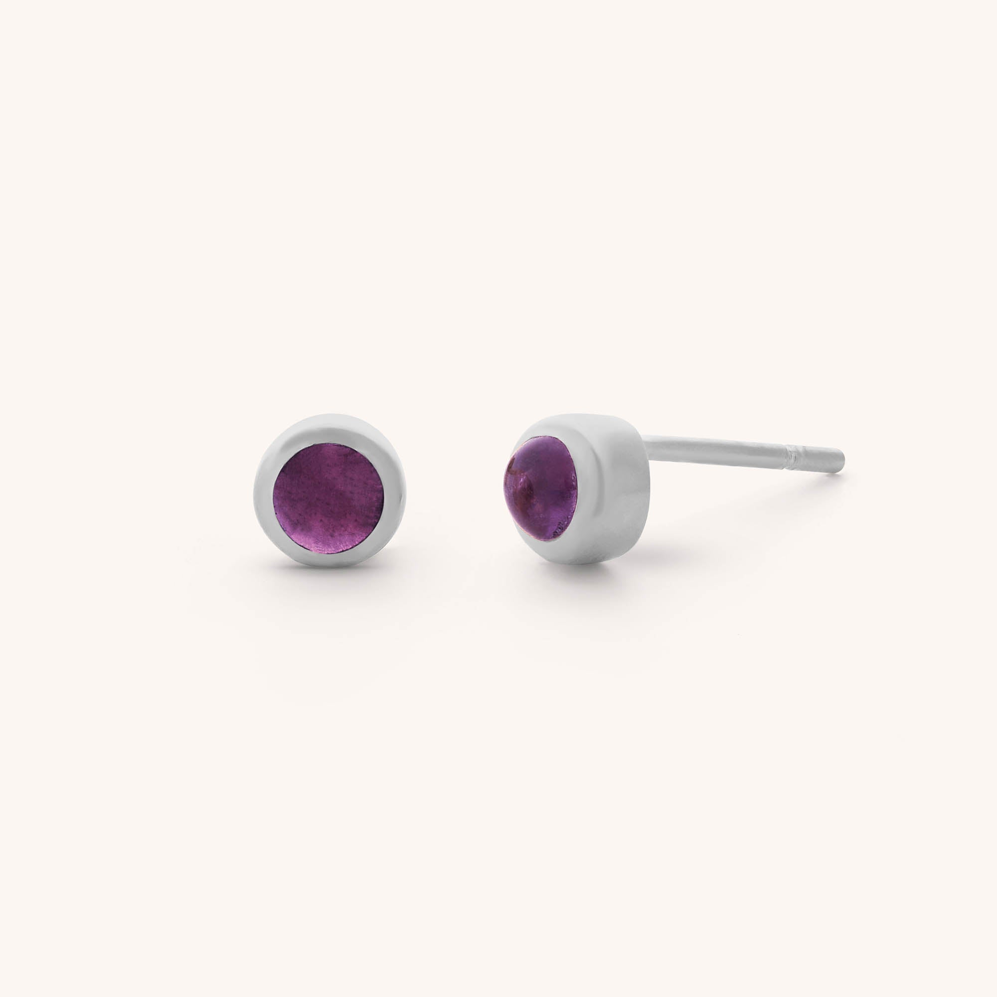 The Mini Gem Dot Birthstone Earrings for February are the perfect gift, featuring round amethyst gemstones set in silver studs. One is shown frontally, and the other from the side, accentuating their simple yet elegant design against a plain background.