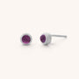 The Mini Gem Dot Birthstone Earrings for February are the perfect gift, featuring round amethyst gemstones set in silver studs. One is shown frontally, and the other from the side, accentuating their simple yet elegant design against a plain background.