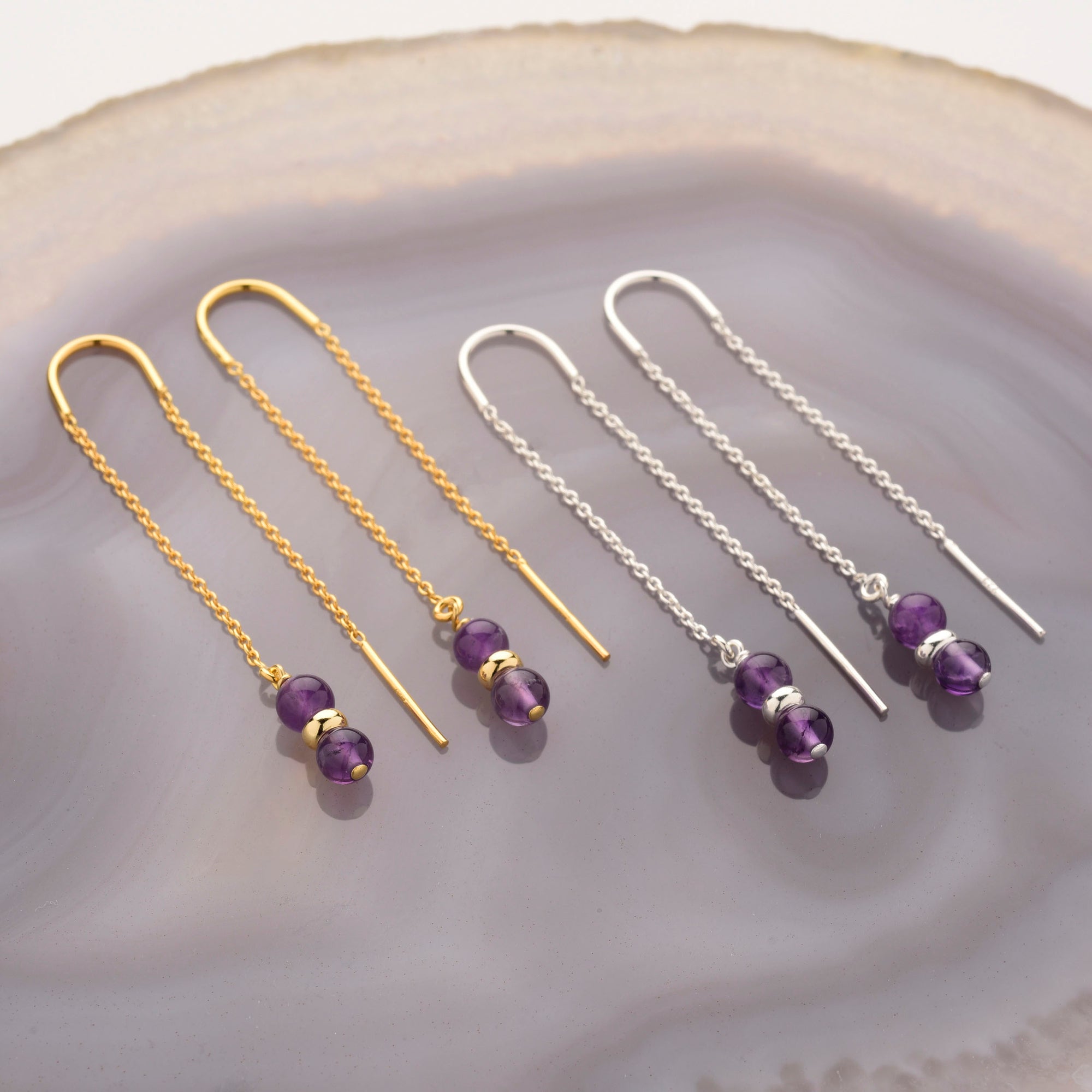 Two pairs of Fall Earrings Birthstone February—threader amethyst earrings with purple gemstone beads and gold or silver chains—are displayed on polished agate, with gold on the left and silver on the right, ideal as a February birthstone gift.