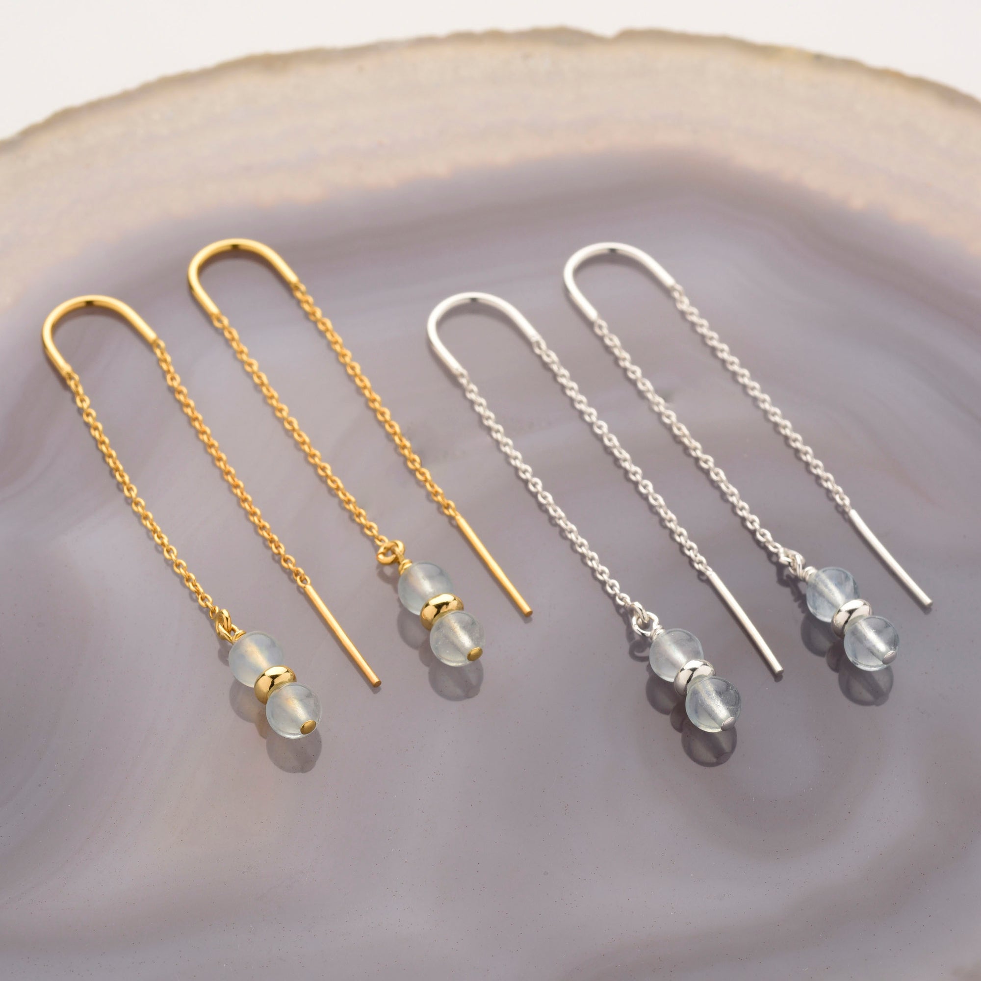 Four Fall Earrings Birthstone March, two in gold and two in sterling silver, each with a delicate chain and two translucent beads, are displayed on a textured, light purple agate surface.