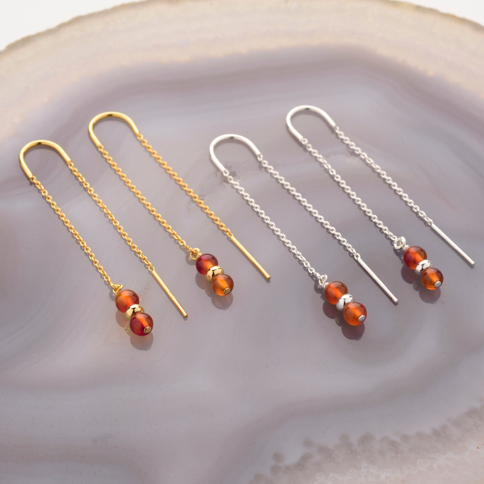 Two pairs of Fall Earrings Birthstone July—threader earrings with chain dangles and three carnelian beads each, one in gold and one in silver—are displayed on a polished agate surface.
