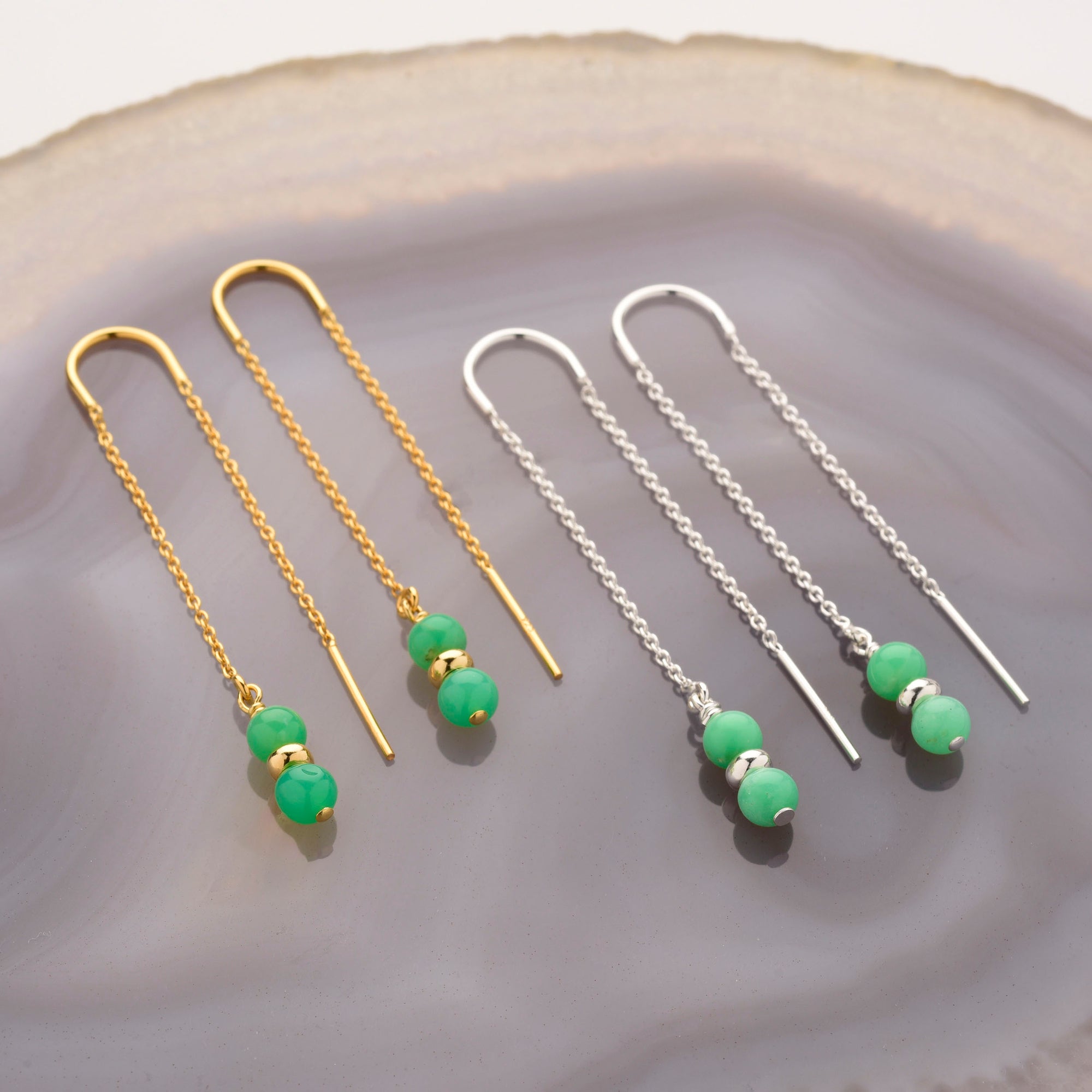 Two pairs of Fall Earrings Birthstone May—dangling chain earrings with green chrysoprase beads, one gold with a plain bar and chain, the other silver—are displayed on a gray marble-style surface.