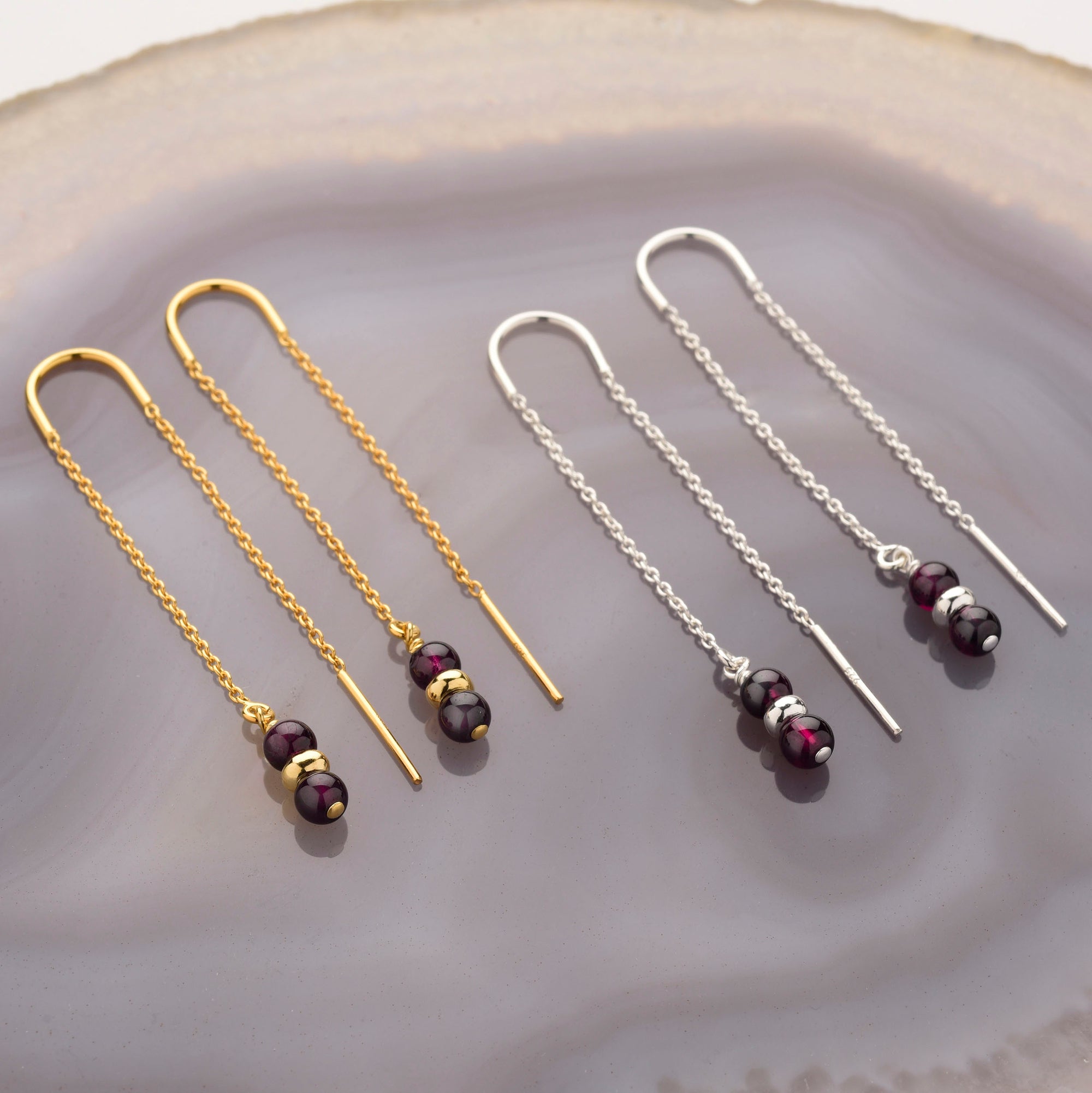 Two pairs of Fall Earrings Birthstone January are shown on polished agate: one gold, one silver, both with dark garnet-like beads and metallic accents at the ends of delicate threader-style chains.
