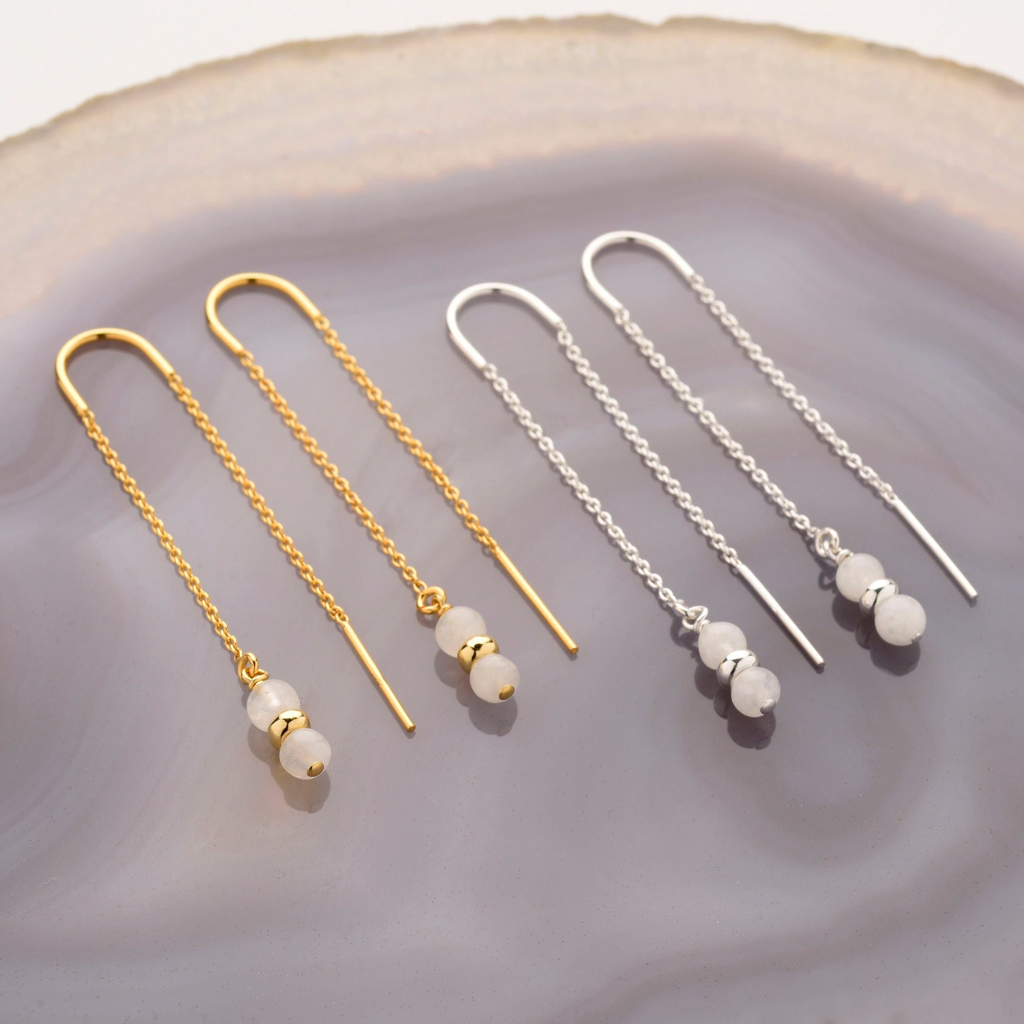 Two pairs of Fall Earrings Birthstone June—one gold, one silver—each with a delicate chain and three small white stone beads, displayed on a smooth light purple agate surface; perfect elegant dangle threader gemstone jewelry.
