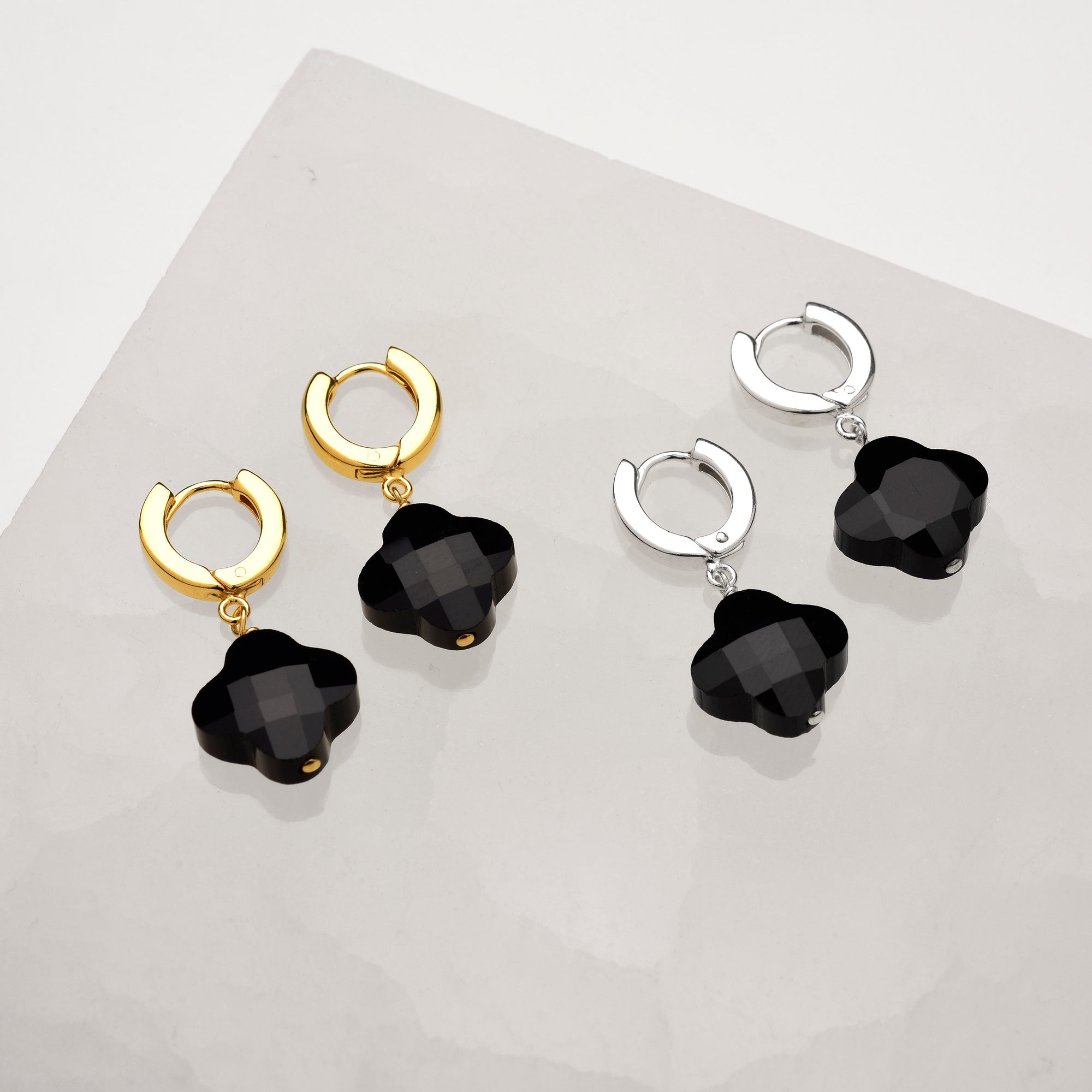 Two pairs of Clover Gem Hoop Earrings—one with gold and one with sterling silver hoops, both featuring black faceted gemstone flower pendants—are displayed on a light, textured surface.