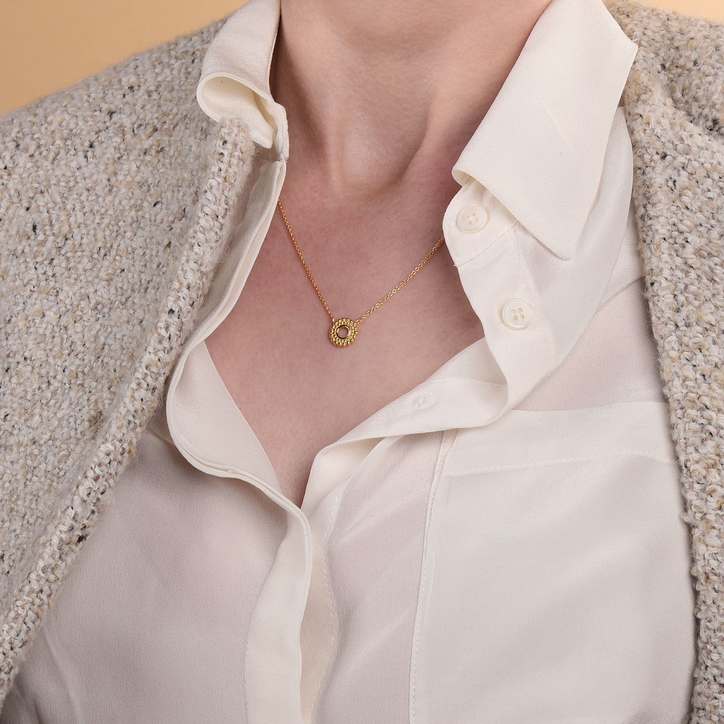 A person in a cream blouse and textured beige jacket showcases the Beaded Halo Necklace, an 18ct gold plated piece with a small circular pendant. The blouse is slightly unbuttoned at the top.