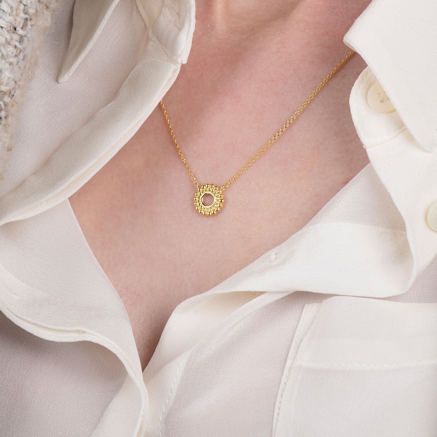 A close-up of a person wearing the Beaded Halo Necklace, crafted in 18ct gold-plated sterling silver with a circular pendant and pink center stone, styled over an open-collar white button-up shirt.