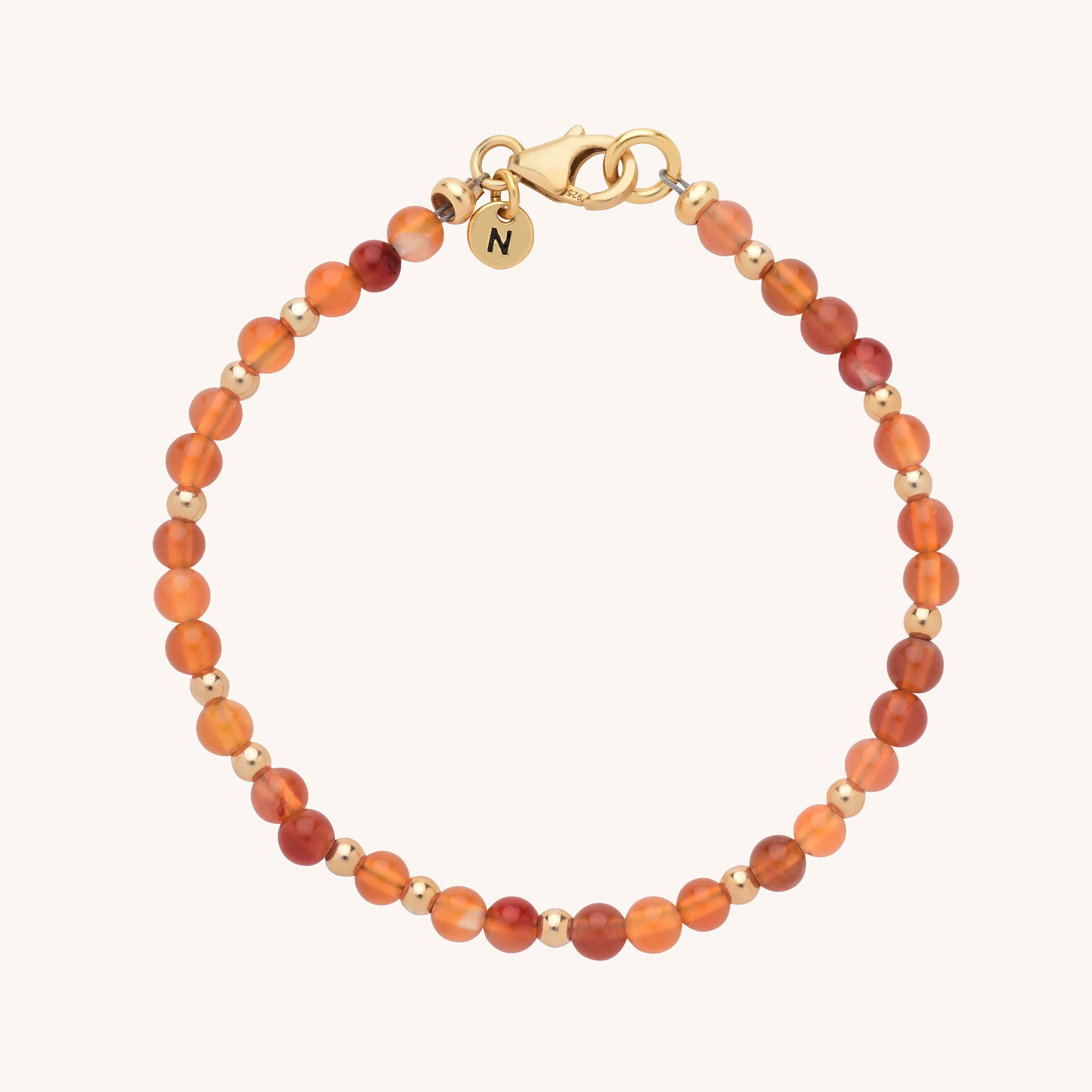 The Jewel Bead Birthstone Bracelet - July features small round carnelian stones, gold beads, a gold lobster clasp, and a gold charm engraved with N, all arranged elegantly on a light background.