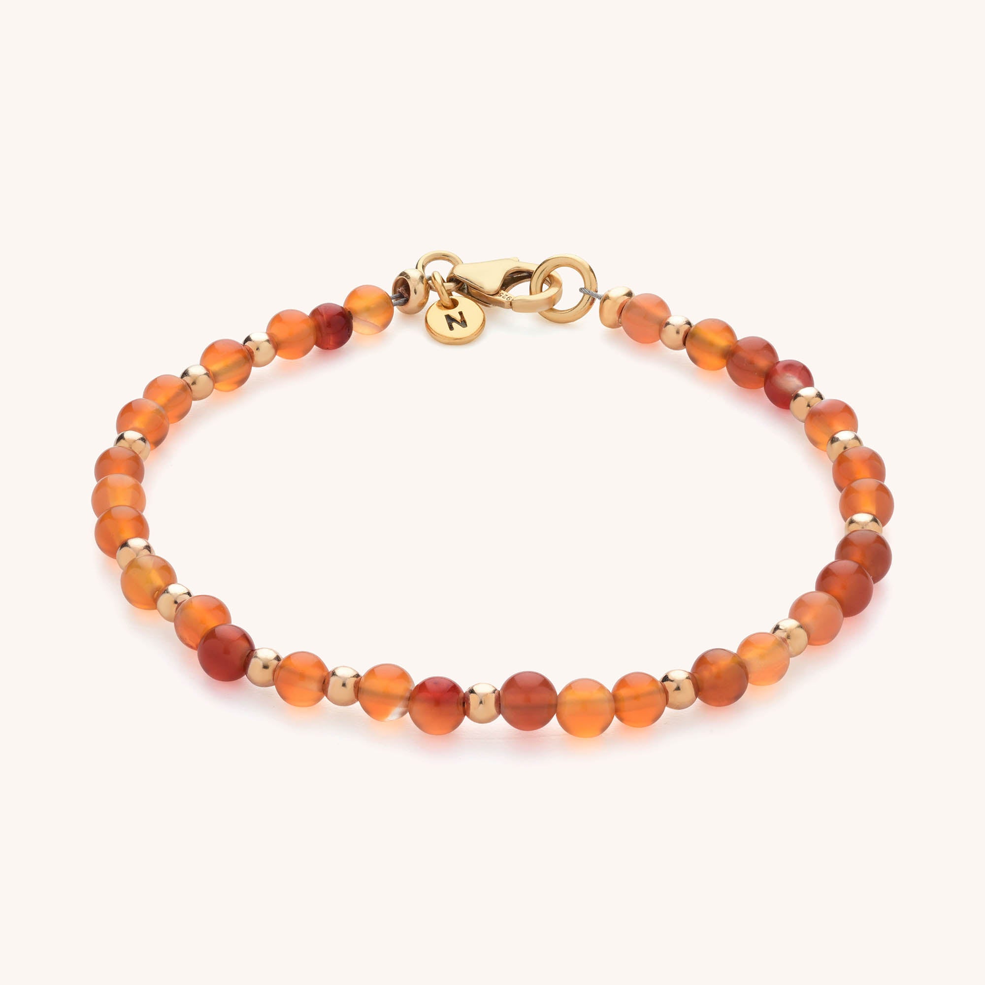 The Jewel Bead Birthstone Bracelet - July features small round carnelian stones, gold spacer beads, a gold lobster clasp, and a round gold charm engraved with the letter N.