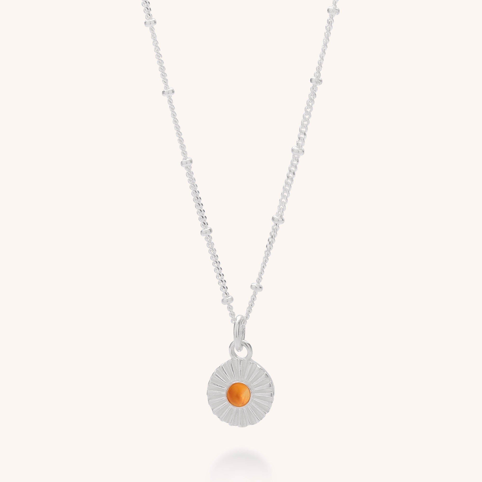 The Radiance Birthstone Necklace features a sterling silver beaded chain and a round sunburst pendant with a vibrant orange semi-precious stone at the center.