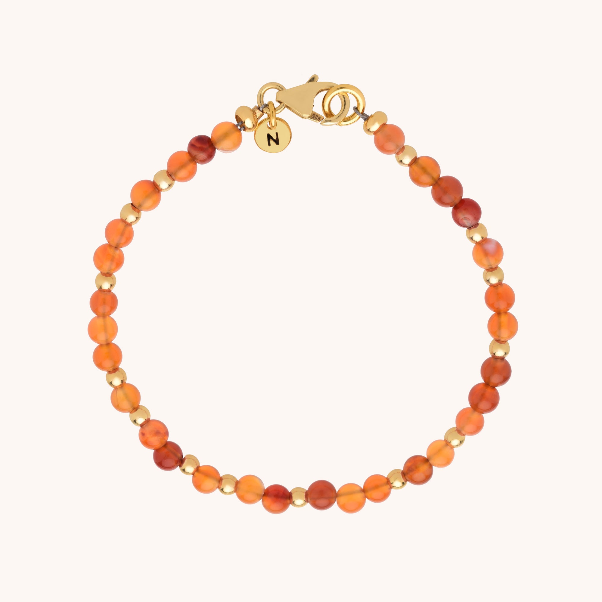 The Jewel Bead Bracelet showcases small round orange and red gemstone beads, gold accents, a gold clasp, and a round gold charm engraved with N for an elegant touch.