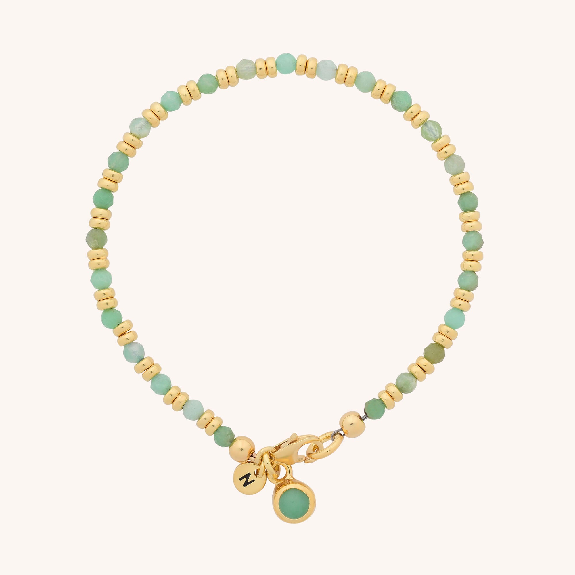 The Linear Birthstone Bracelet - May features alternating gold and chrysoprase-green beads, a gold clasp, and two charms: a round gold tag with “N” and a green stone pendant—an ideal gift for May birthdays.