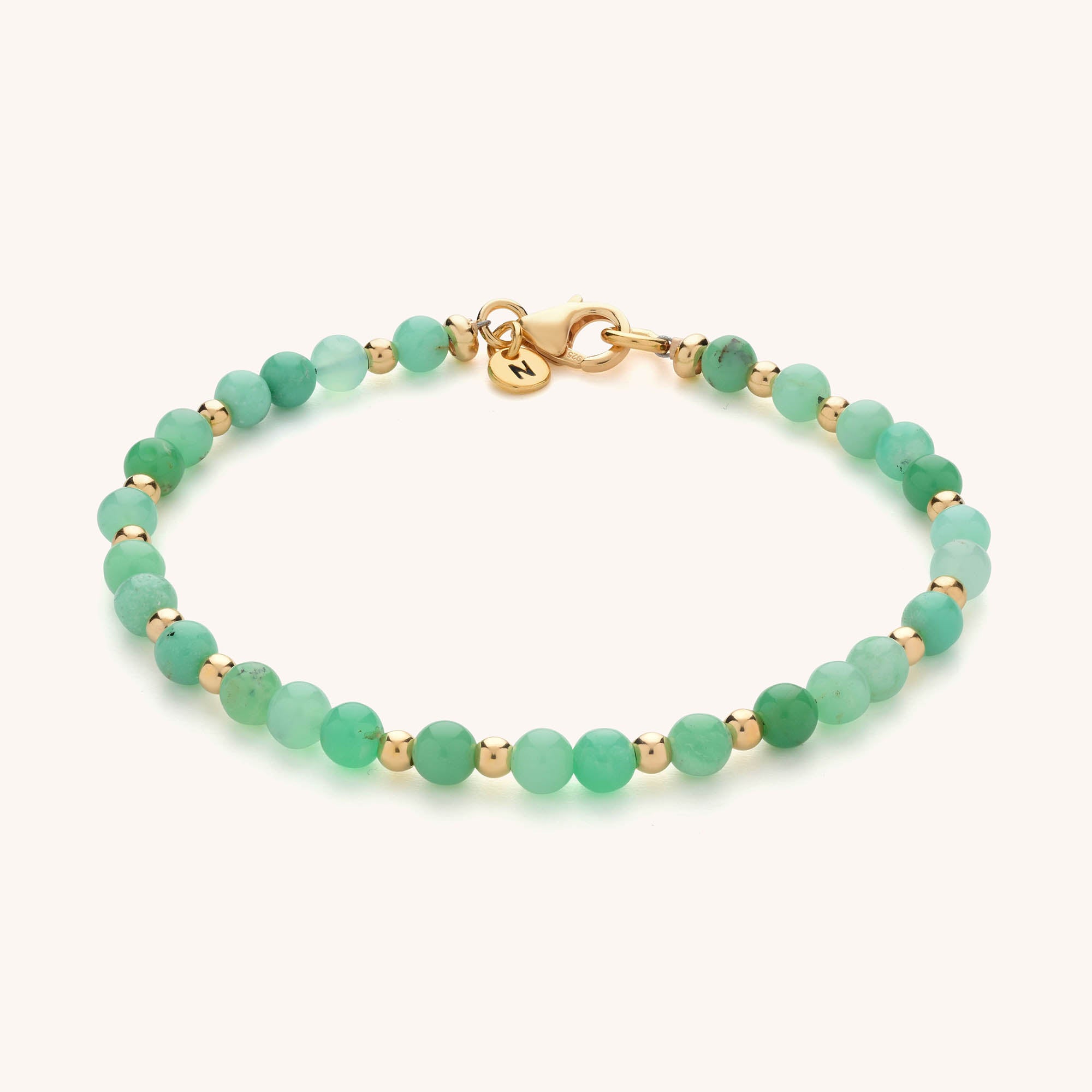 The Jewel Bead Birthstone Bracelet features round green beads, small gold spacers, a gold clasp, and a round gold tag with an engraved N. An elegant birthstone bracelet perfect for adding a personalized touch.
