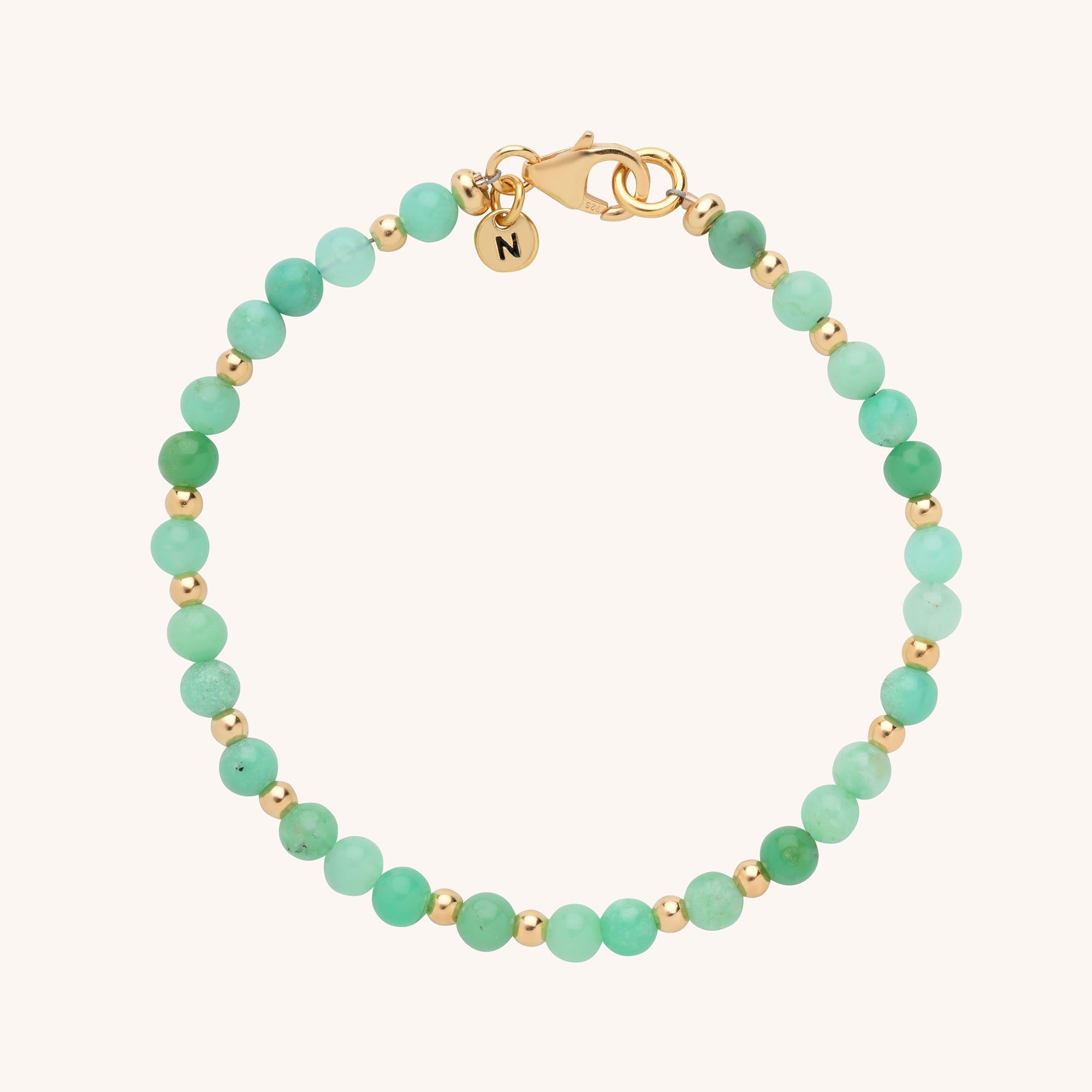 The Jewel Bead Birthstone Bracelet features alternating small green and gold beads, a gold lobster clasp, and a round charm engraved with N—an elegant, personalized accessory for any look.